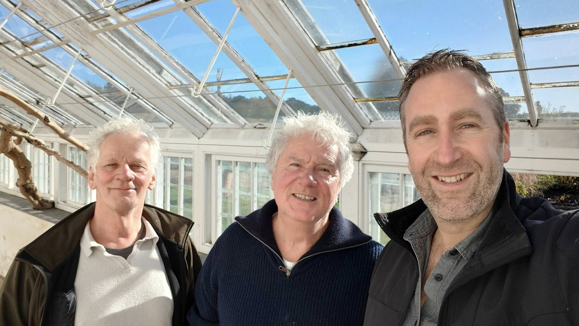 Drie mannen poseren samen in een kas met glazen plafond en muren; ze lachen naar de camera.