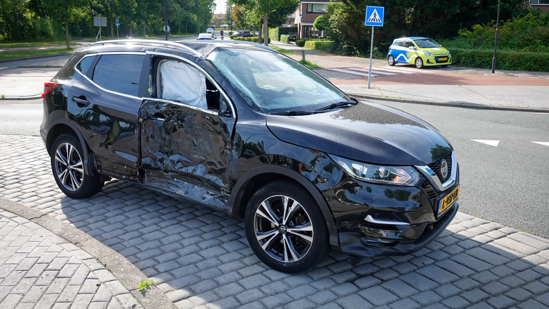 Zwarte auto met aanzienlijke schade aan de zijkant, geparkeerd op een straat met bestrating en een zebrapad op de achtergrond.