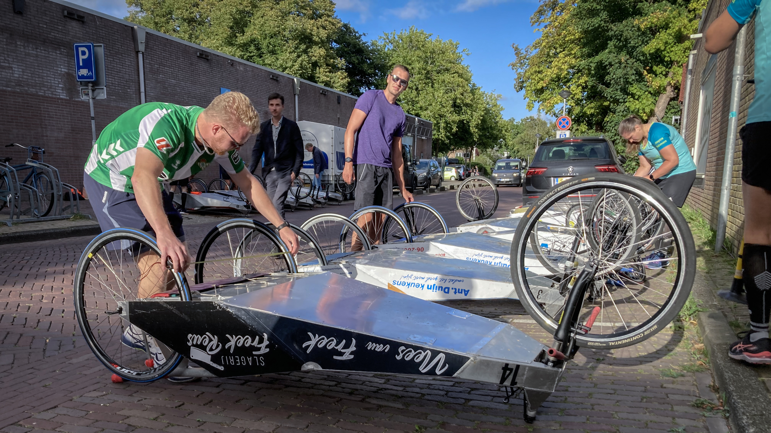 Een groep mensen op straat werkt aan ligfietsen, waarschijnlijk voor een race of evenement. Ze monteren of repareren de fietsen terwijl ze zich in een rij bevinden. De straat heeft aan de zijkant geparkeerde auto's en bomen op de achtergrond.