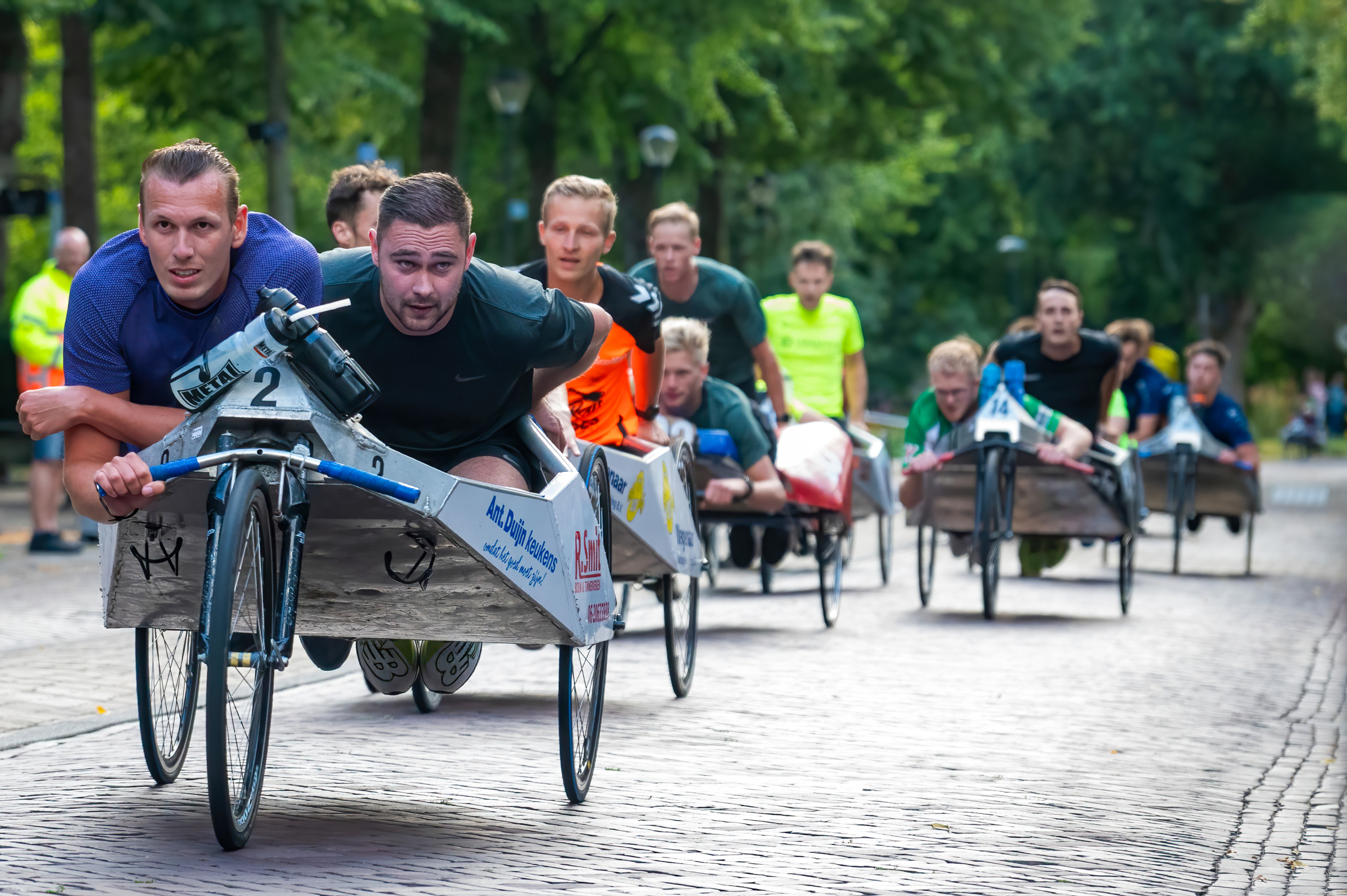 Mannen nemen deel aan een race in zelfgemaakte voertuigen op drie wielen, liggend op houten platforms, op een straat met bomen op de achtergrond.