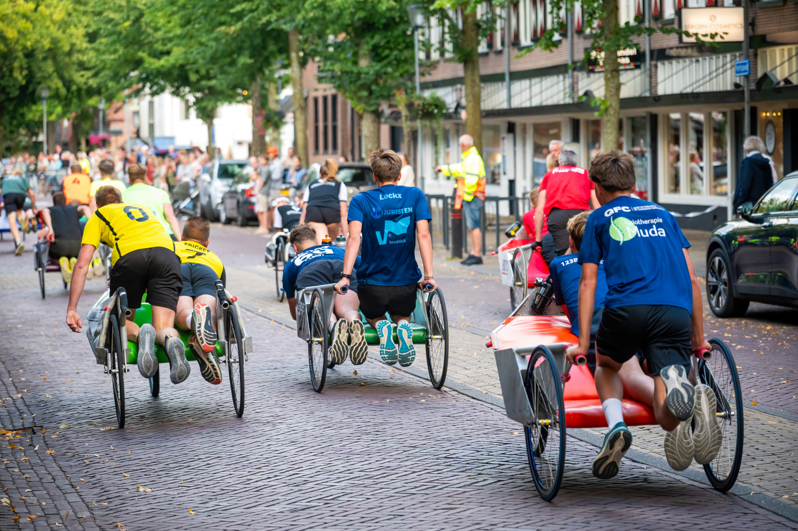 Mensen nemen deel aan een ludieke race met fietsen en ligstoelen op een geplaveide straat, terwijl toeschouwers langs de kant staan te kijken.