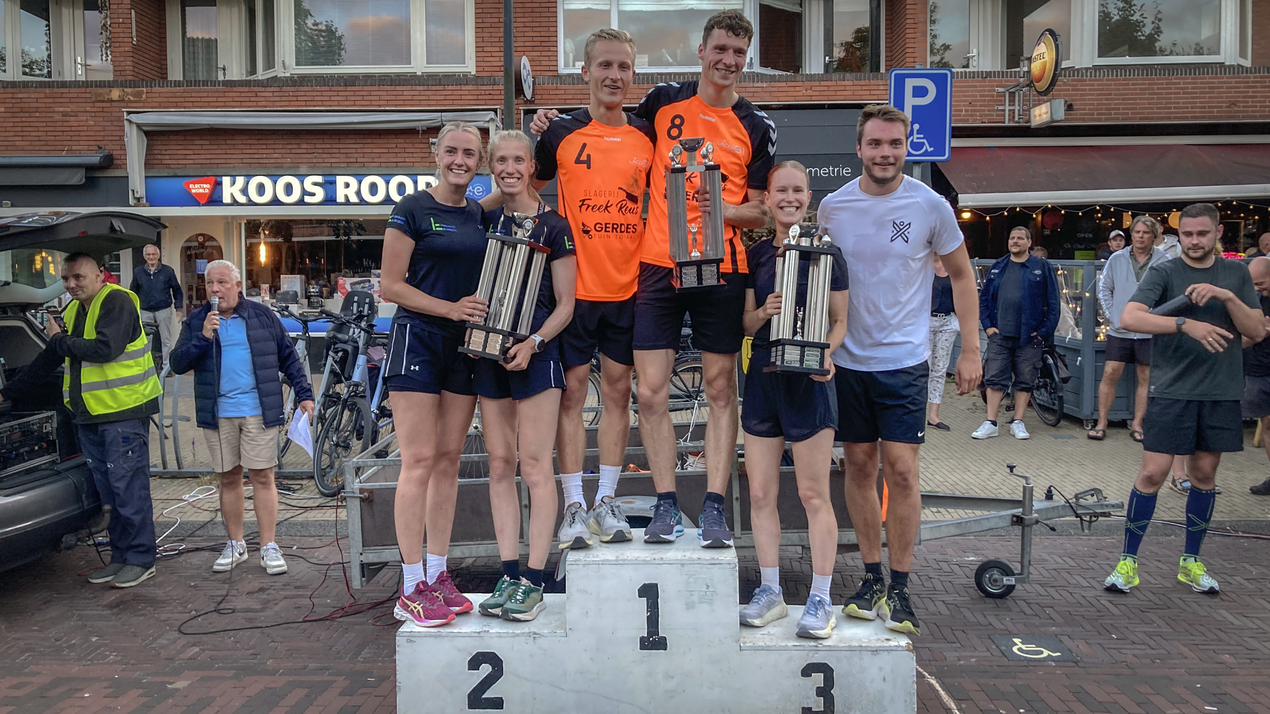 Een groep van zes winnaars staat op een podium, elk met een grote trofee, voor een gebouw met de naam 'Koos Roos'. Drie van hen dragen oranje sportshirts met nummers 4 en 8, de overige drie dragen donkerblauwe sportkleding. Op de achtergrond staan toeschouwers en is een man met een microfoon bezig met de aankondiging.