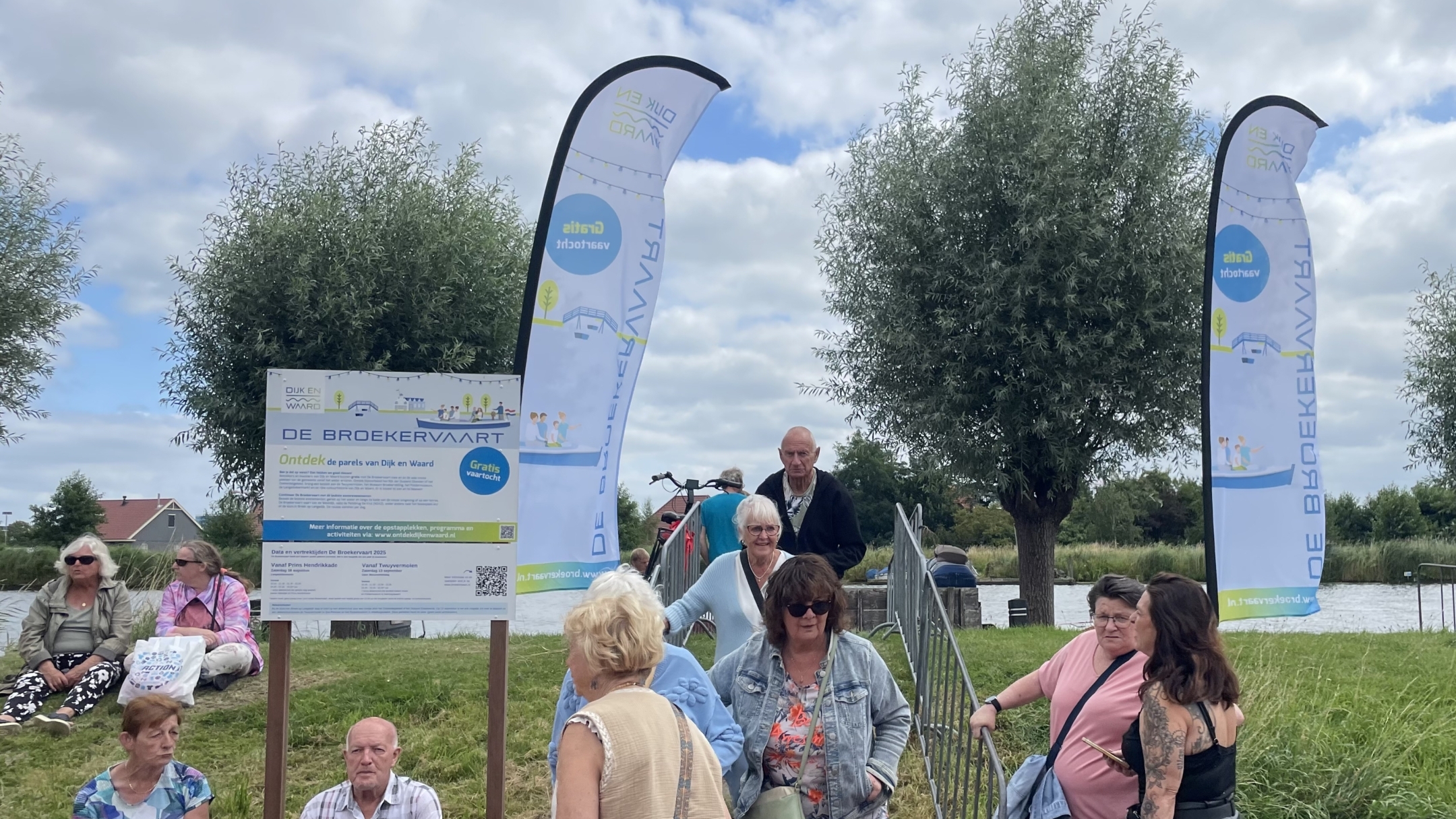 Groep mensen rond een reclamebord en vlaggen met de tekst 'De Broekervaart' bij een grasveld met bomen en een water op de achtergrond, op een bewolkte dag.