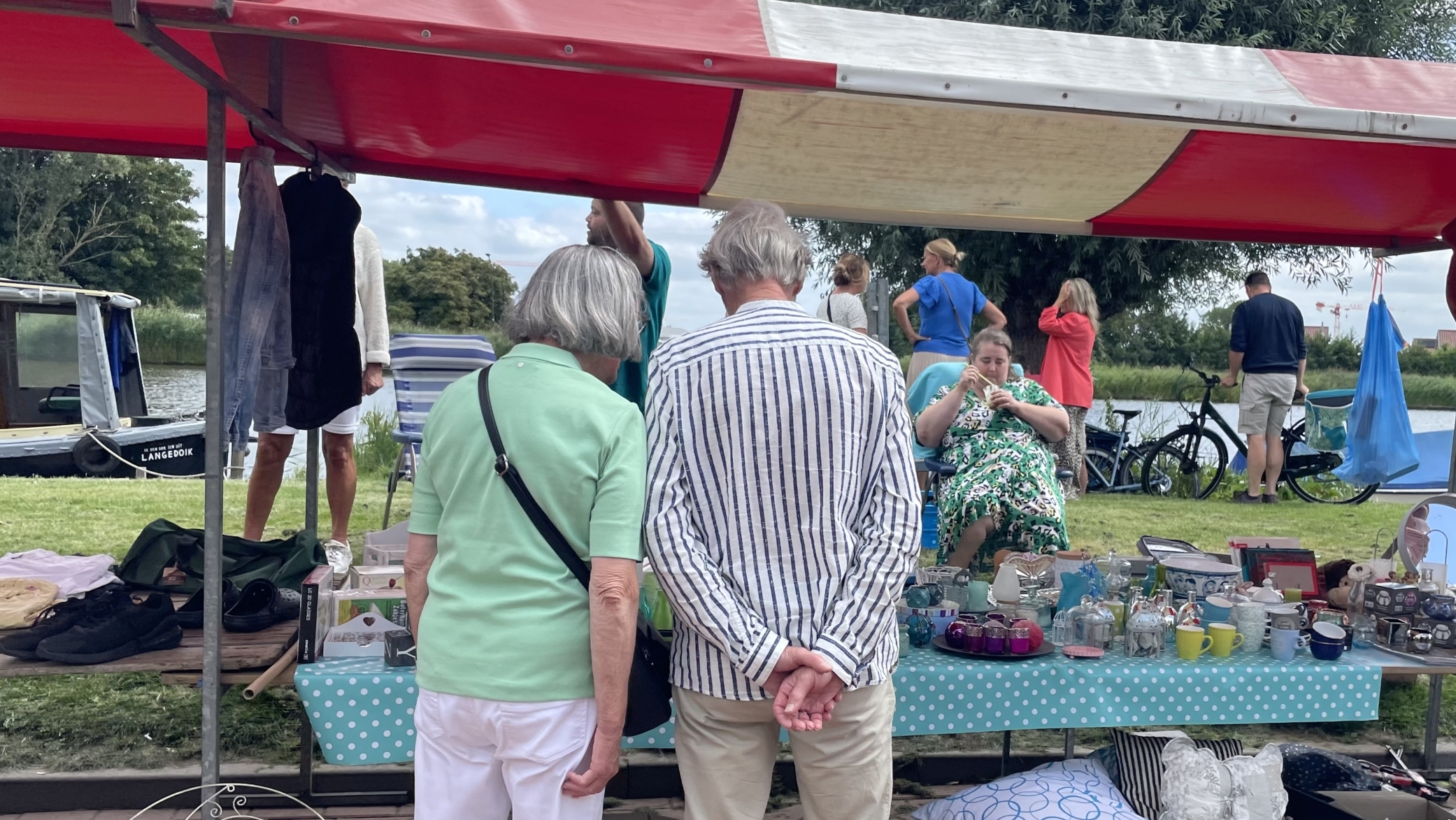 Onder een rode en witte marktkraam staan mensen op een rommelmarkt, met op de tafel diverse tweedehands spullen zoals kopjes en glazen. Op de achtergrond zijn een boot en fietsen te zien in een parkachtige omgeving.