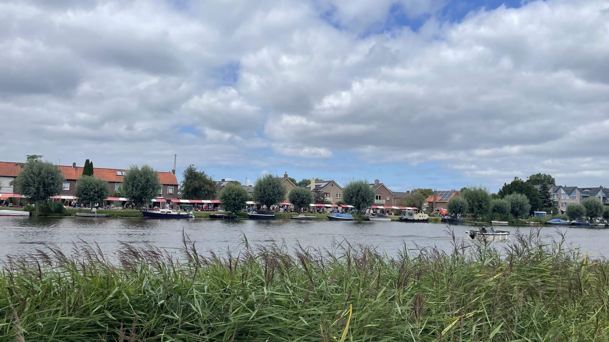 Uitzicht op een rivier met op de achtergrond huizen met rode daken en groene bomen, met boten aangemeerd langs de oever en riet in de voorgrond onder een bewolkte hemel.