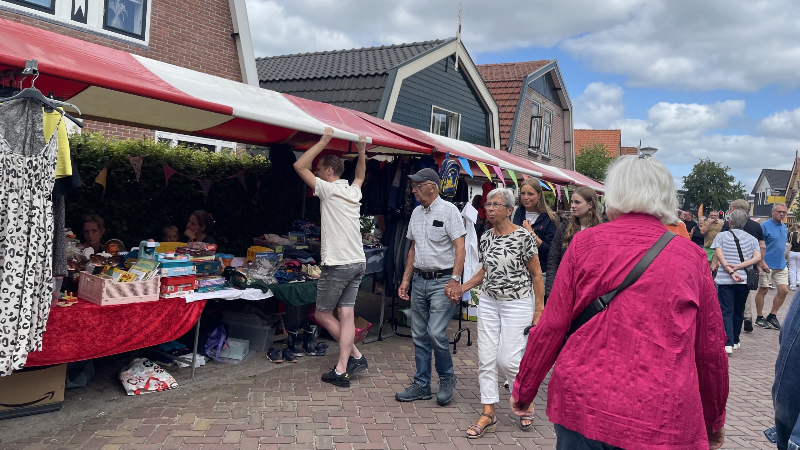 Personen lopen langs een kraam op een straatmarkt met diverse spullen uitgestald, waaronder kleding en speelgoed, onder een rood-wit gestreept afdak.