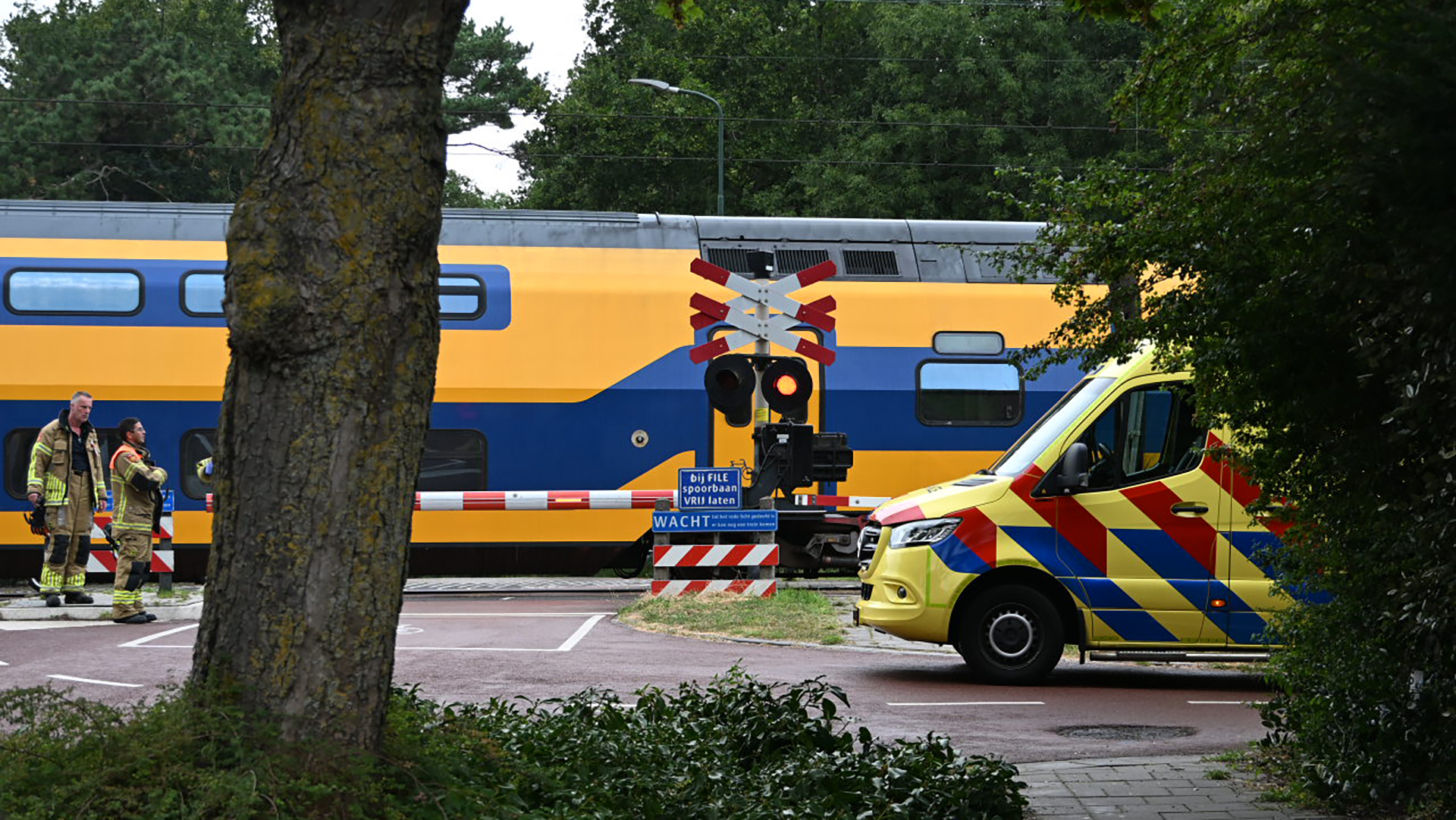 Een stilstaande trein bij een spoorwegovergang met een gele en blauwe Nederlandse ambulance geparkeerd aan de zijkant, omgeven door bomen en struiken, en twee brandweerlieden die op de achtergrond staan.