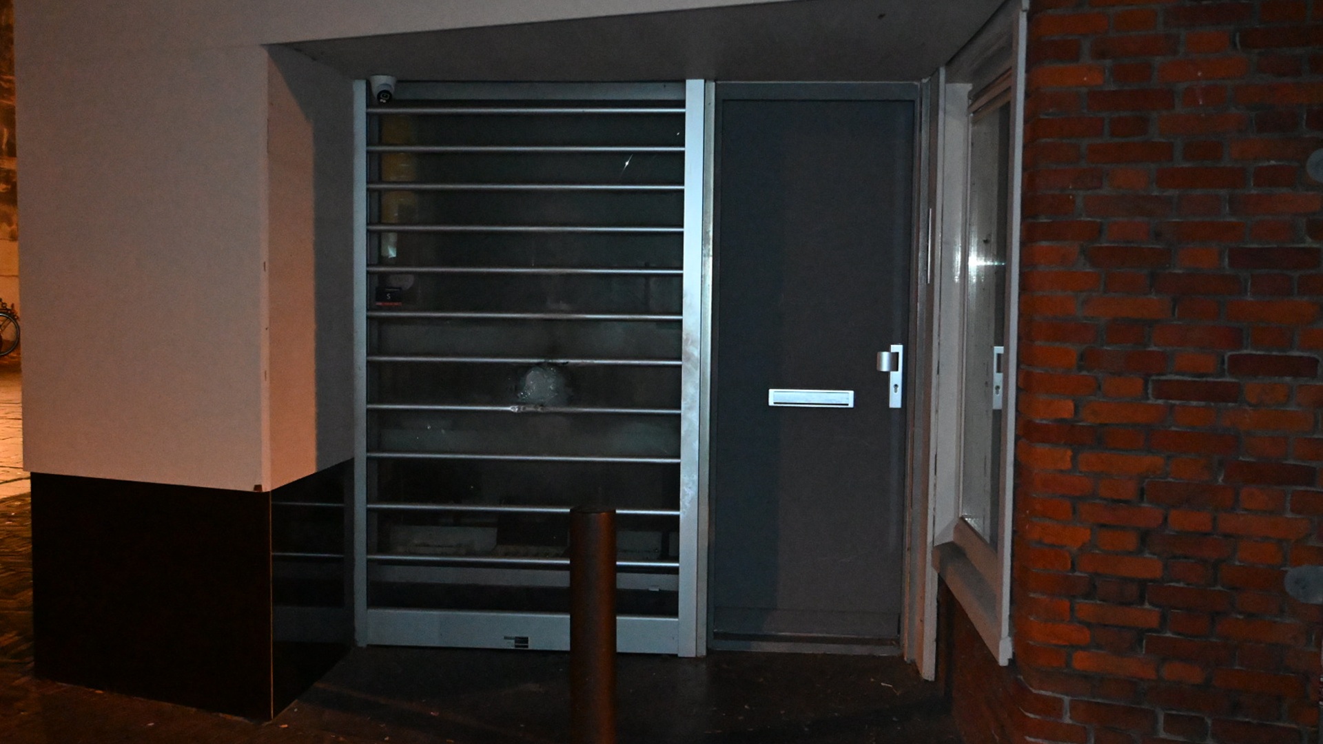 Een metalen hek en grijze deur naast elkaar in een hoek van een gebouw, met een bakstenen muur aan de rechterkant, gefotografeerd bij nacht.