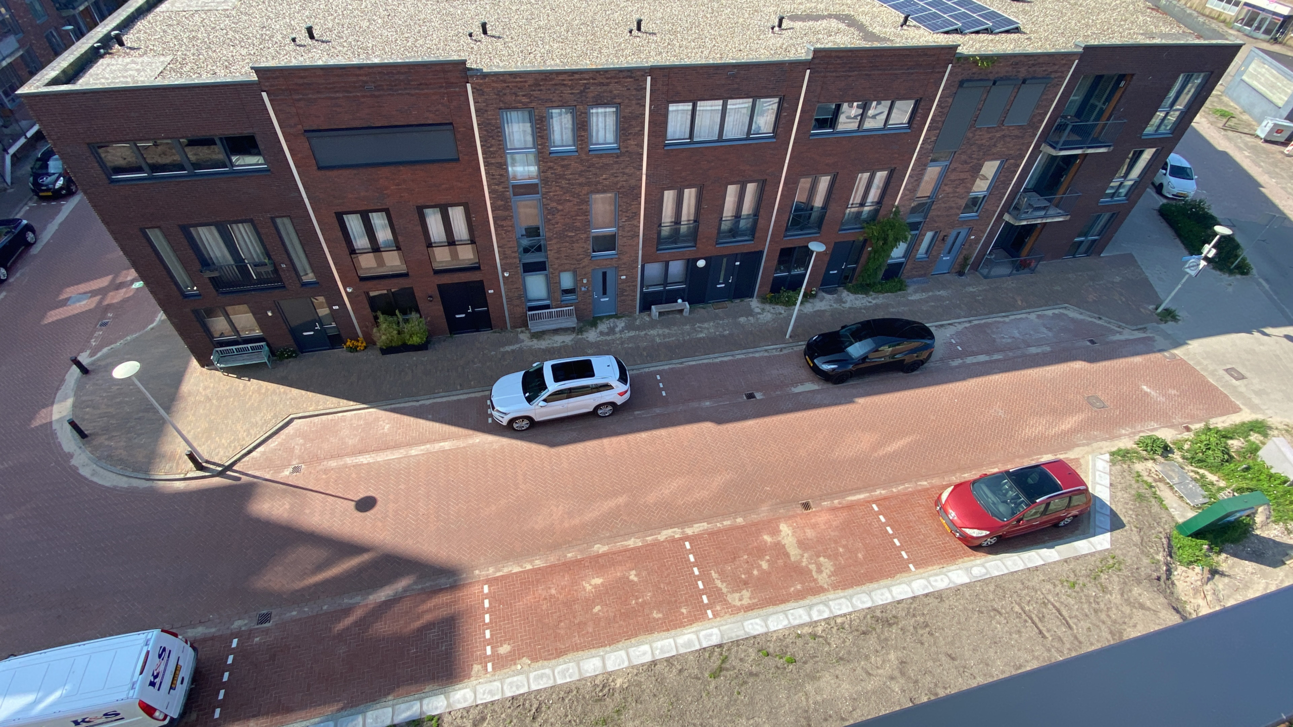 Luchtfoto van een rij moderne bakstenen woningen langs een straat met drie geparkeerde auto's; twee witte auto's en één rode auto.