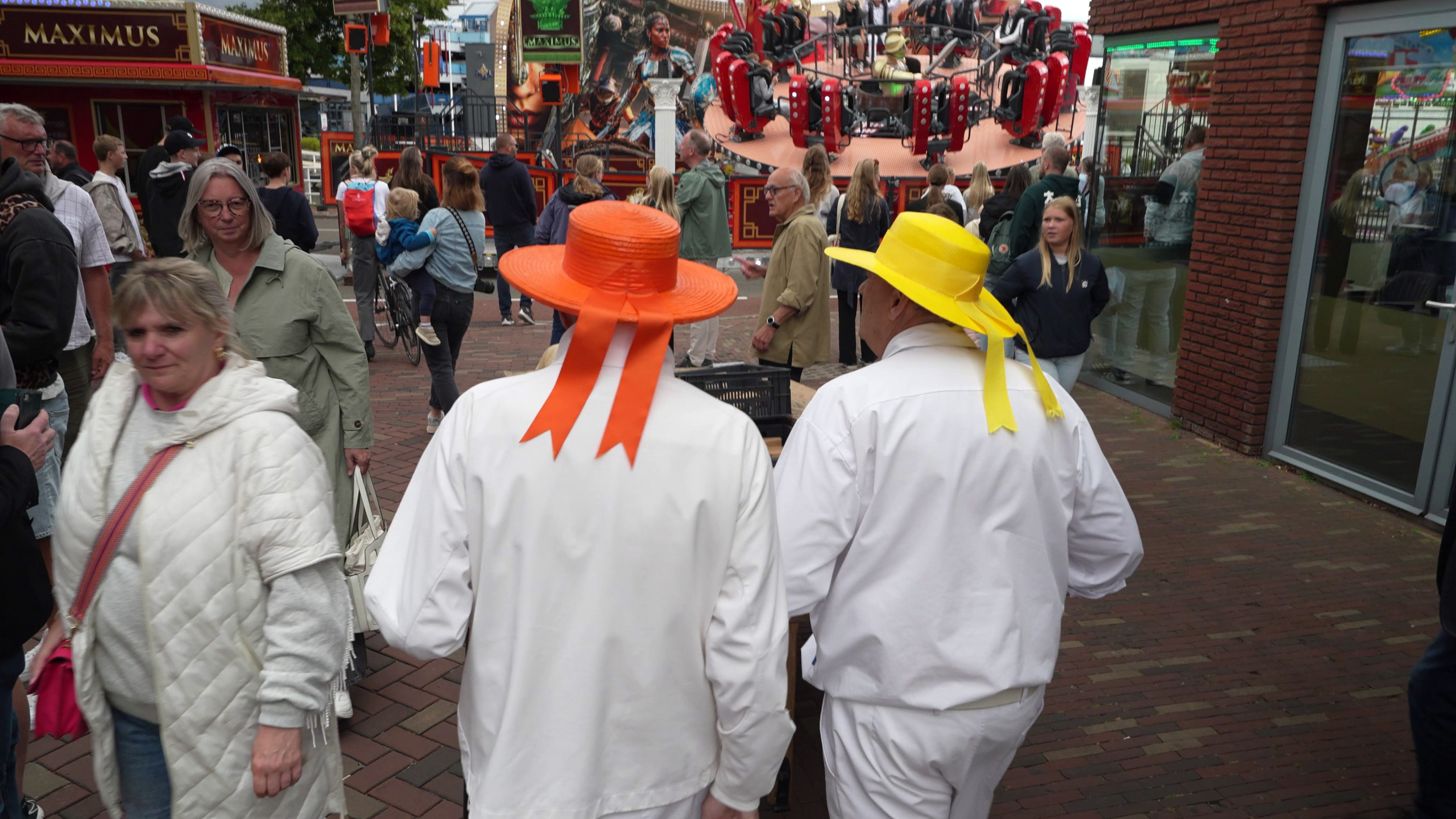 Twee mannen in witte kleding met oranje en gele hoeden lopen hand in hand op een kermis met mensen om hen heen en een attractie op de achtergrond.