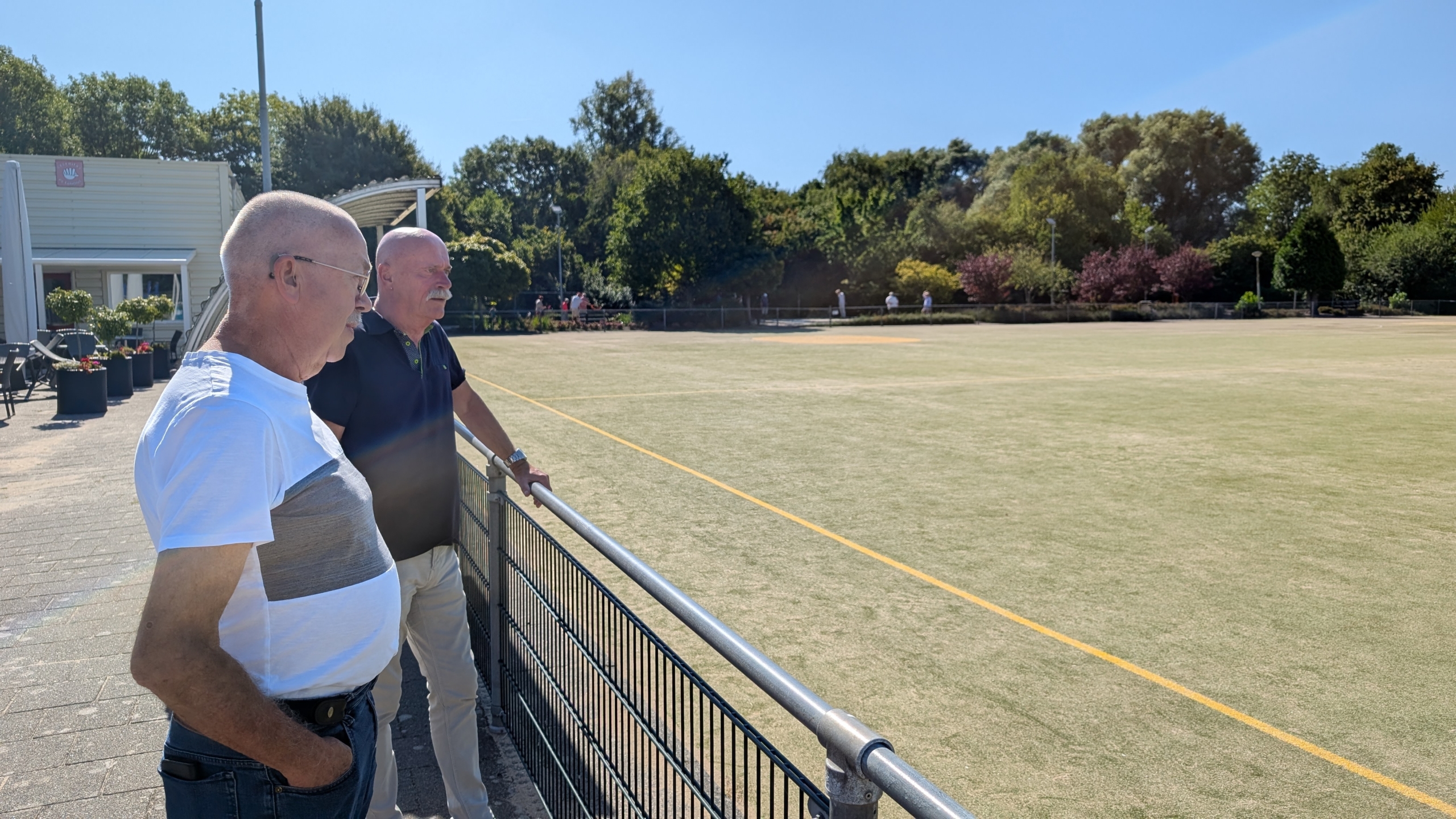 Twee mannen staan bij een hek dat uitkijkt over een grasveld op een zonnige dag, met bomen en struiken op de achtergrond.