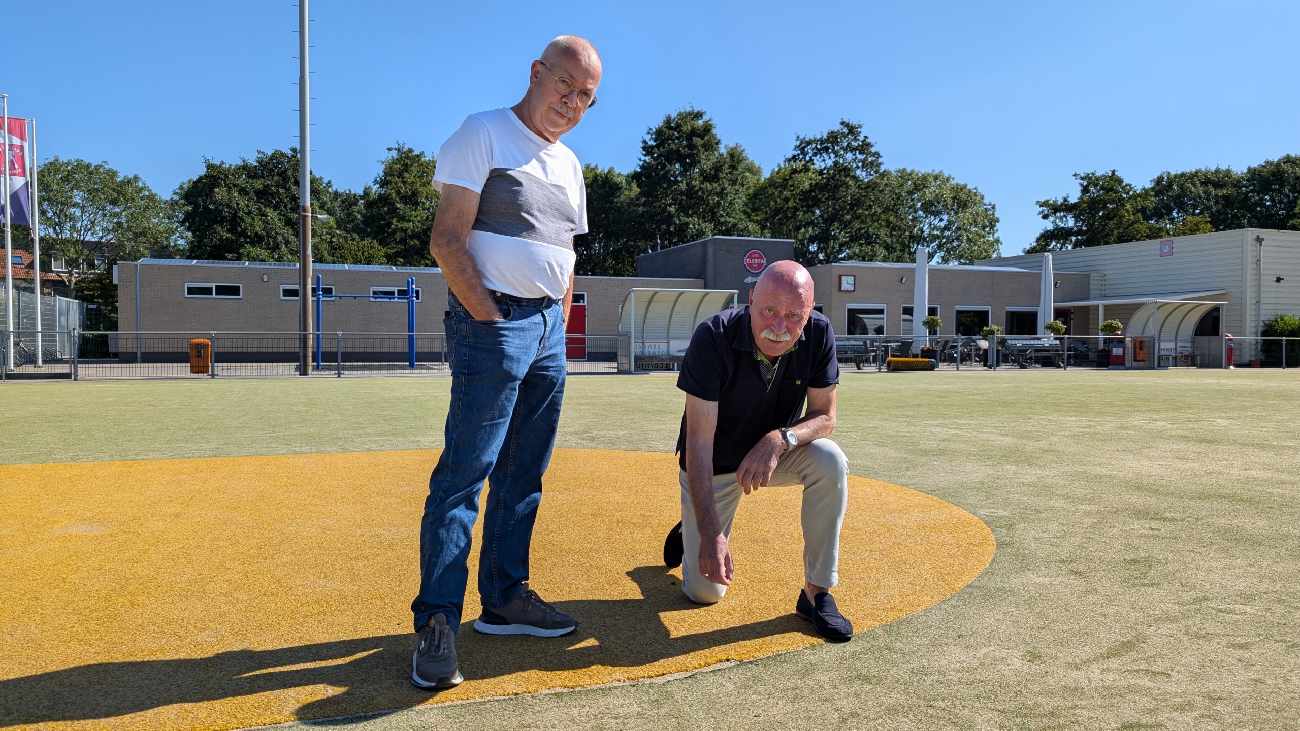 Twee mannen op een sportveld, de ene staat terwijl de andere gehurkt is, met een gebouw en blauwe lucht op de achtergrond.