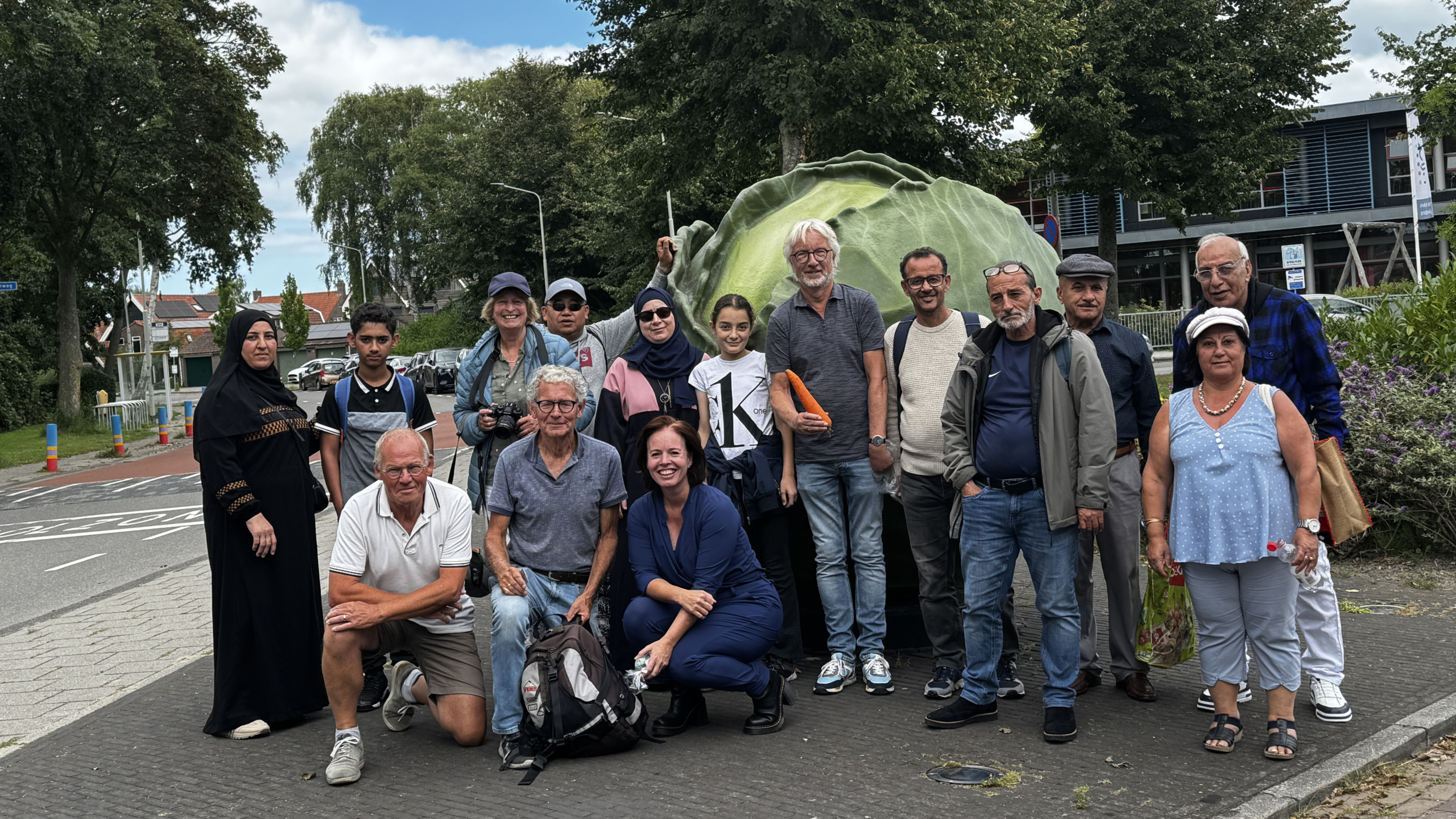 Groep van vijftien mensen poseert samen buiten op een trottoir voor een grote kunstmatige kool, omgeven door bomen en een woonwijk.