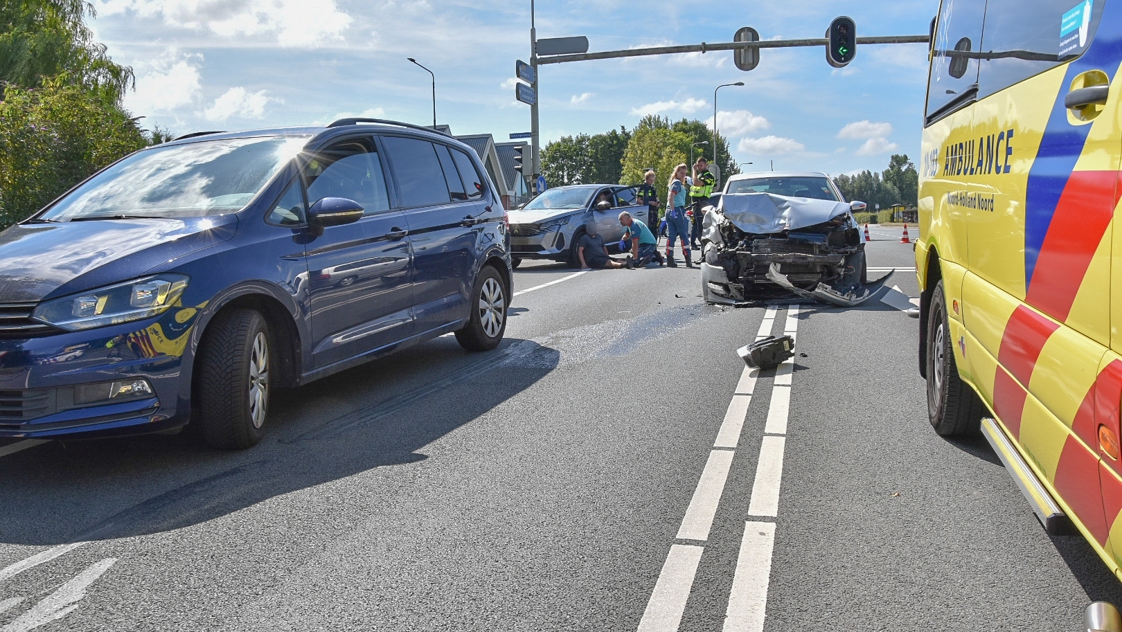 Verkeersongeluk met meerdere auto's, waarbij een voertuig aanzienlijke schade aan de voorzijde heeft; hulpverleners en een gele ambulance zijn aanwezig bij het stoplicht.