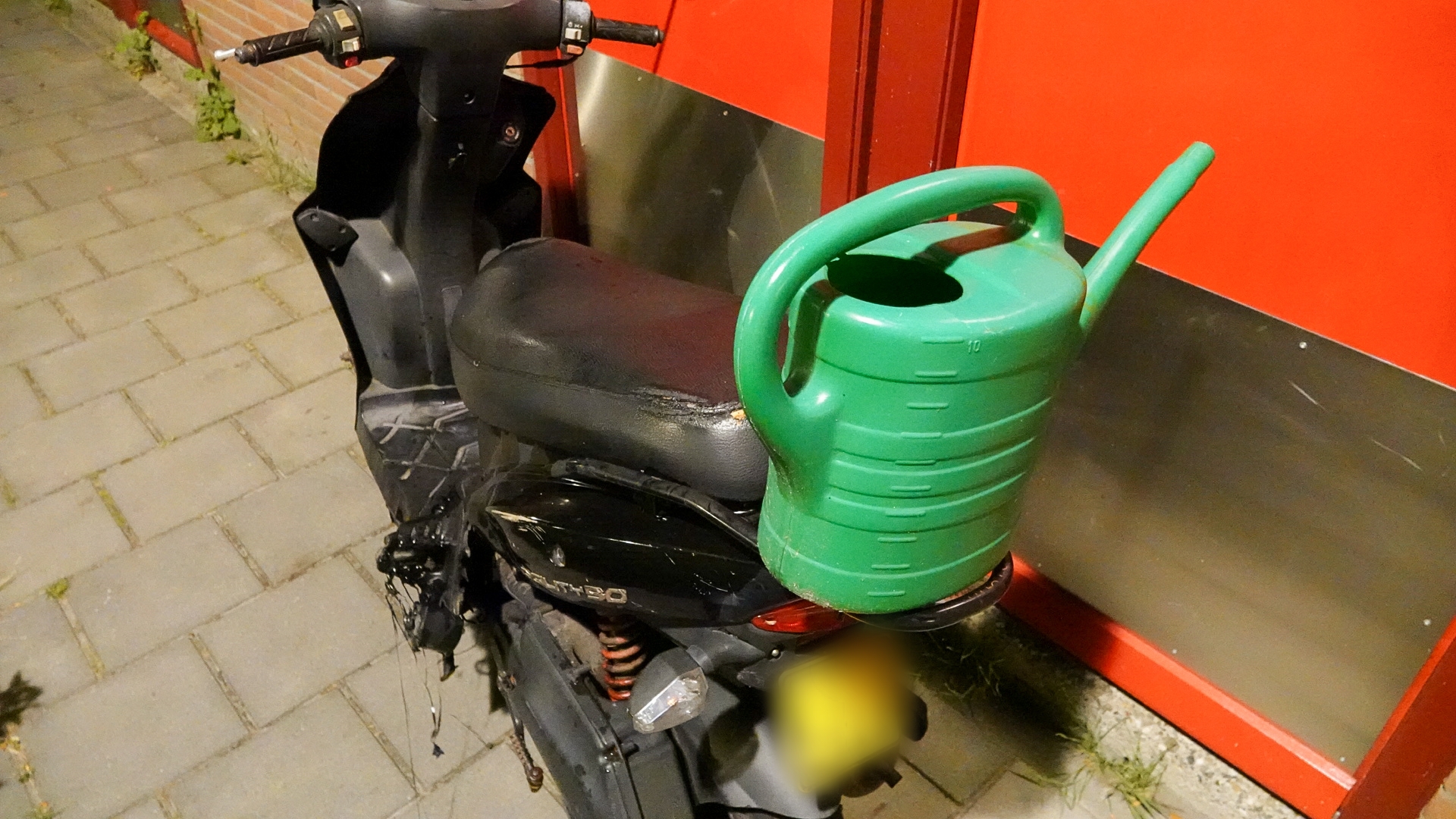 Een groene gieter staat op de zwarte stoel van een scooter, geparkeerd langs een stoep met tegels."