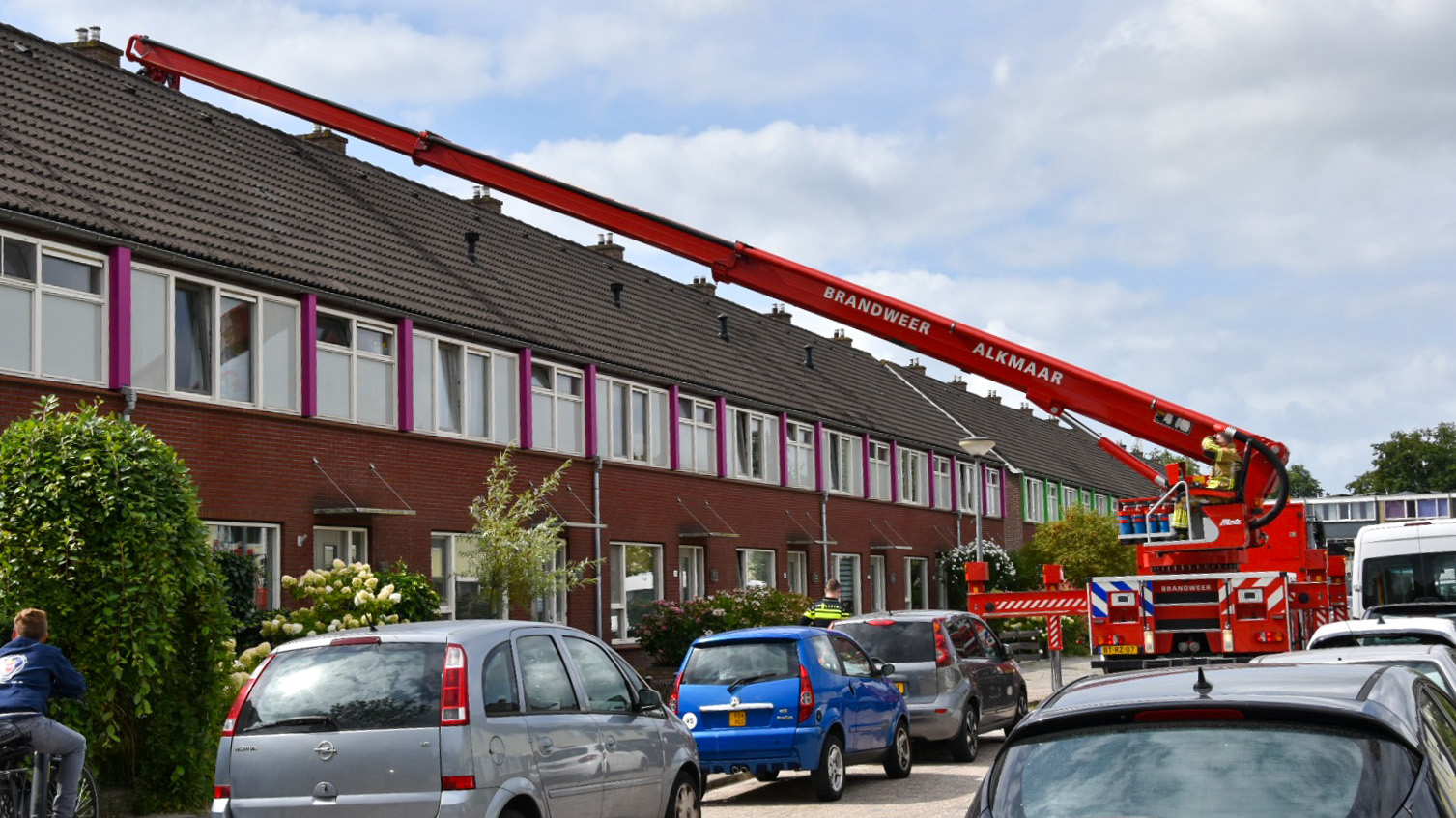 Een brandweerwagen met een lange rode ladder van de brandweer Alkmaar, geparkeerd voor een rijtjeshuis. Een brandweerman staat op het platform van de ladder die naar het dak van het huis reikt. Er staan auto's geparkeerd langs de straat en een kind op een fiets kijkt naar de actie.