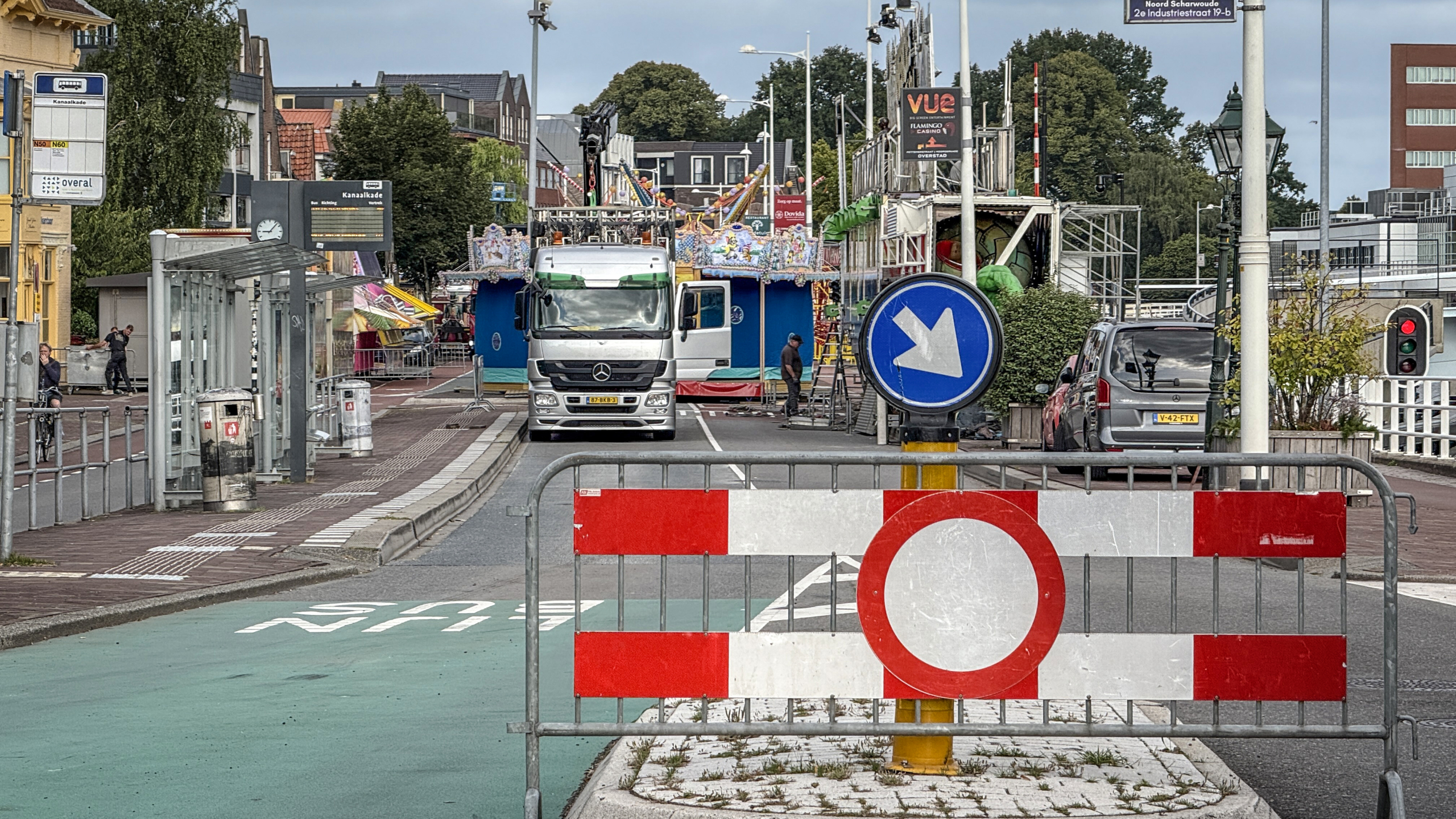 Straat in een stedelijke omgeving, geblokkeerd door een verkeersbord met een rode cirkel en een blauw bord dat naar rechts wijst. Op de achtergrond staat een vrachtwagen voor een kermisattractie, met mensen die bezig zijn met opbouw. Aan de linkerkant is een bushalte zichtbaar.
