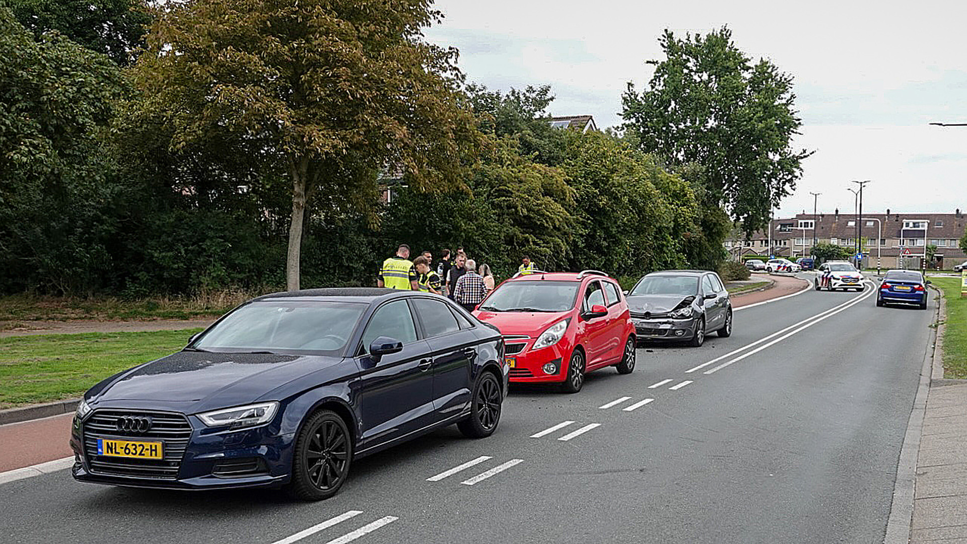 Een verkeersongeval met vier auto's op een weg, waarbij mensen en politieagenten aan de kant met elkaar praten, en een auto zichtbare schade aan de voorkant heeft.