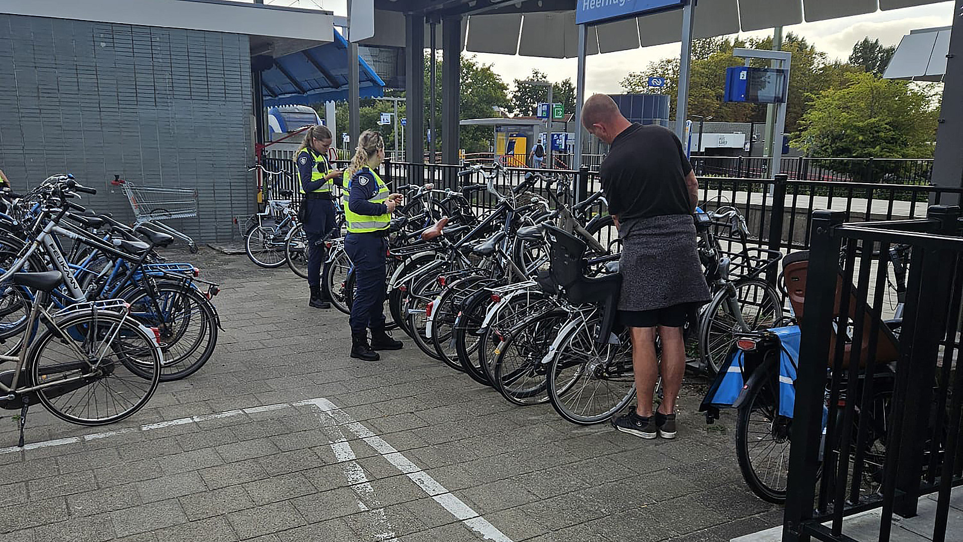 Twee vrouwen in uniform controleren fietsen bij een stalling naast een stationsgebouw, waar ook een man bezig is met een fiets.