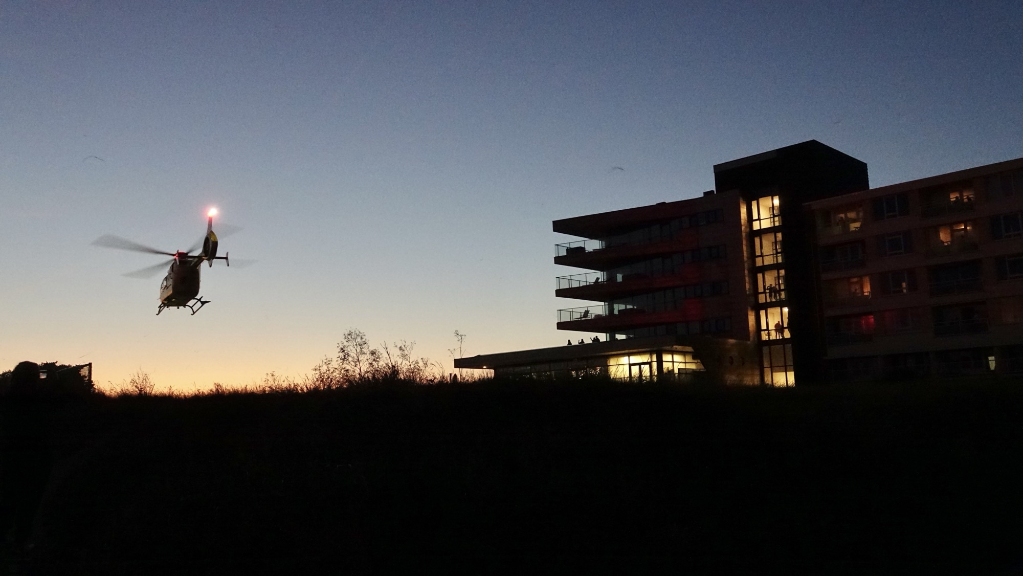 Een helikopter vliegt tijdens de schemering in de buurt van een flatgebouw met verlichte ramen.
