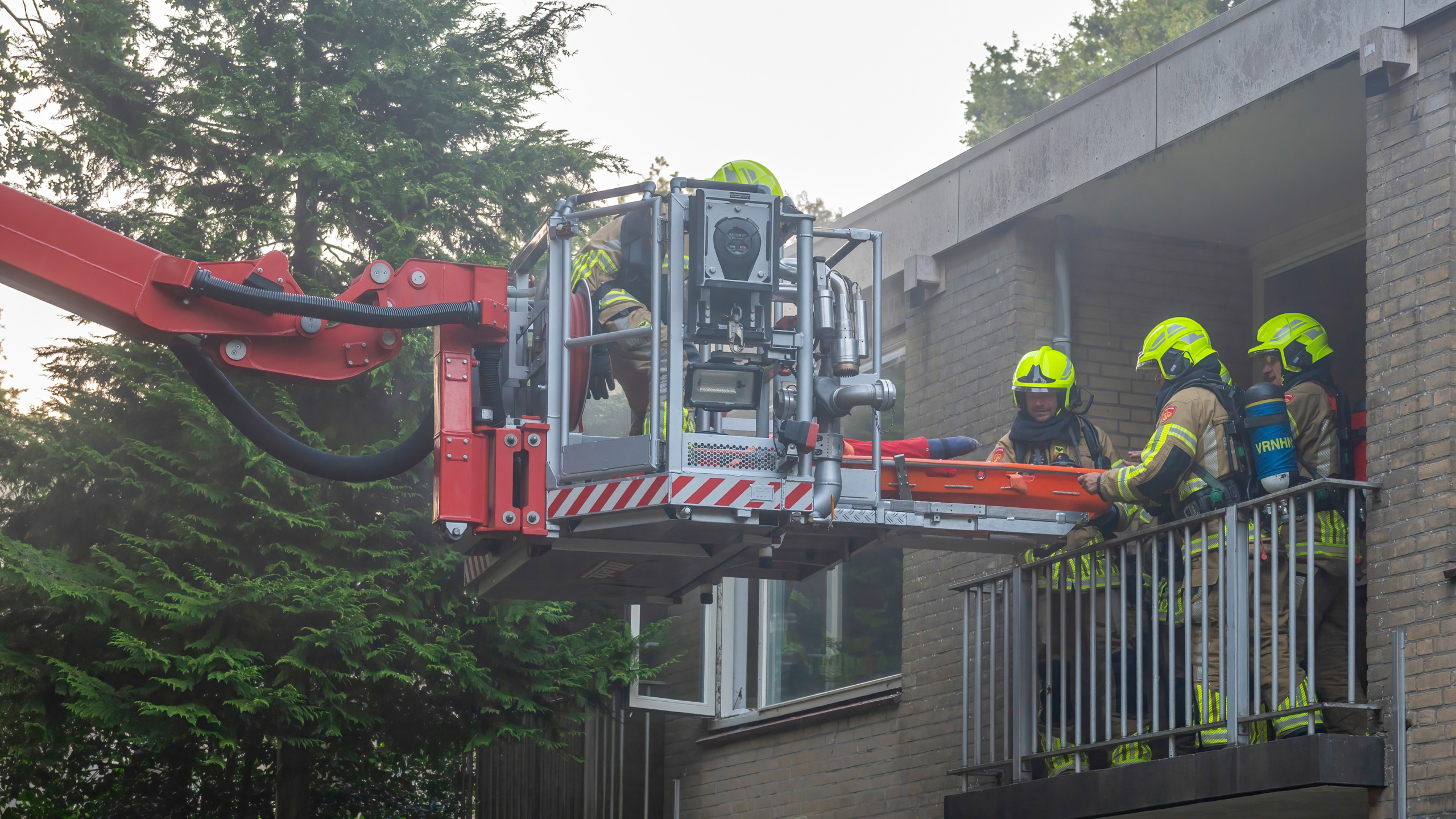 Brandweerlieden in gele helmen en beschermende kleding tillen een brancard naar een platform met een hijskraan aan de zijkant van een gebouw.