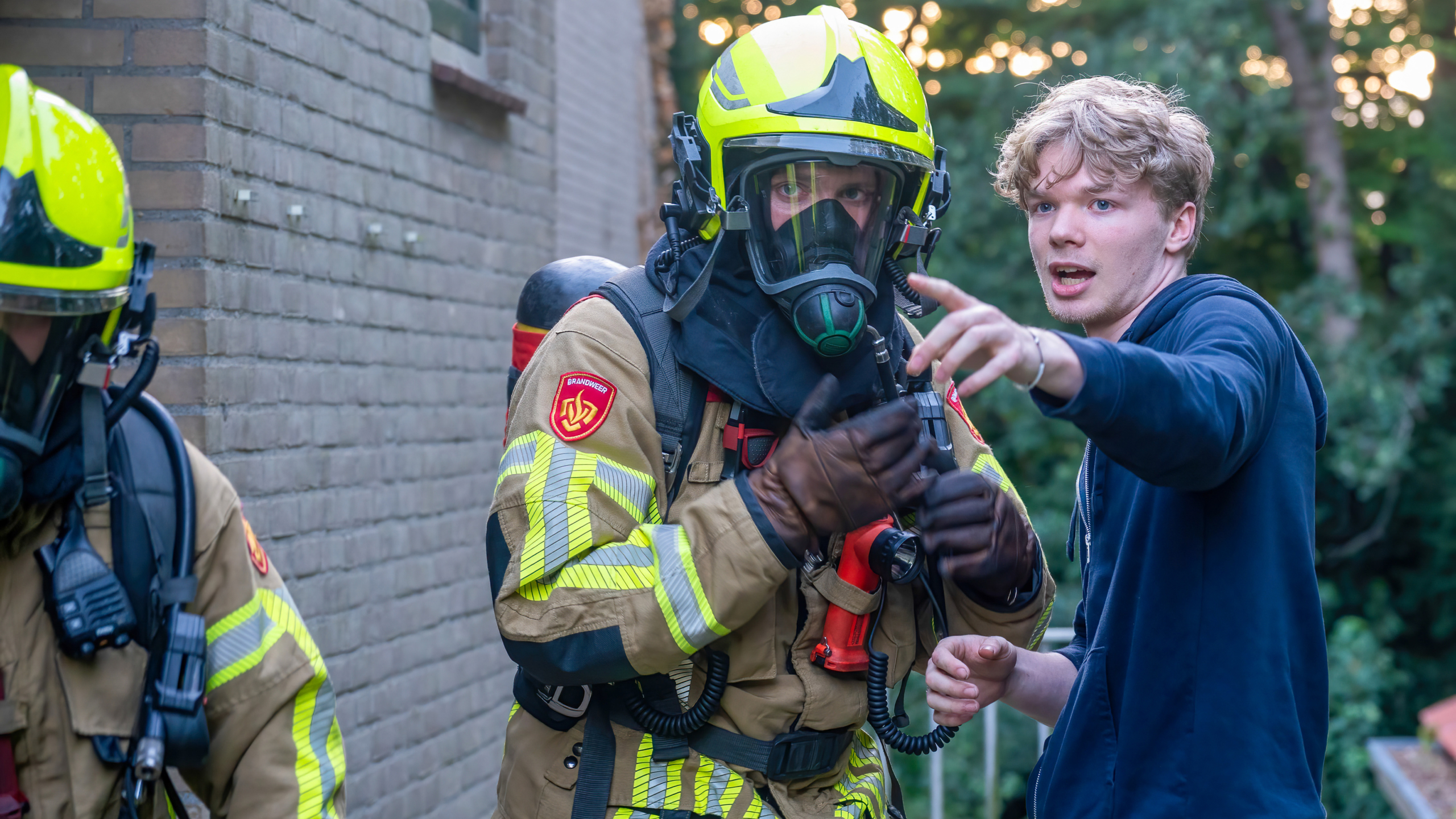 Een brandweerman in volle bescherming met gasmasker en helm staat buiten een gebouw, terwijl een man naast hem wijst richting iets in de verte.