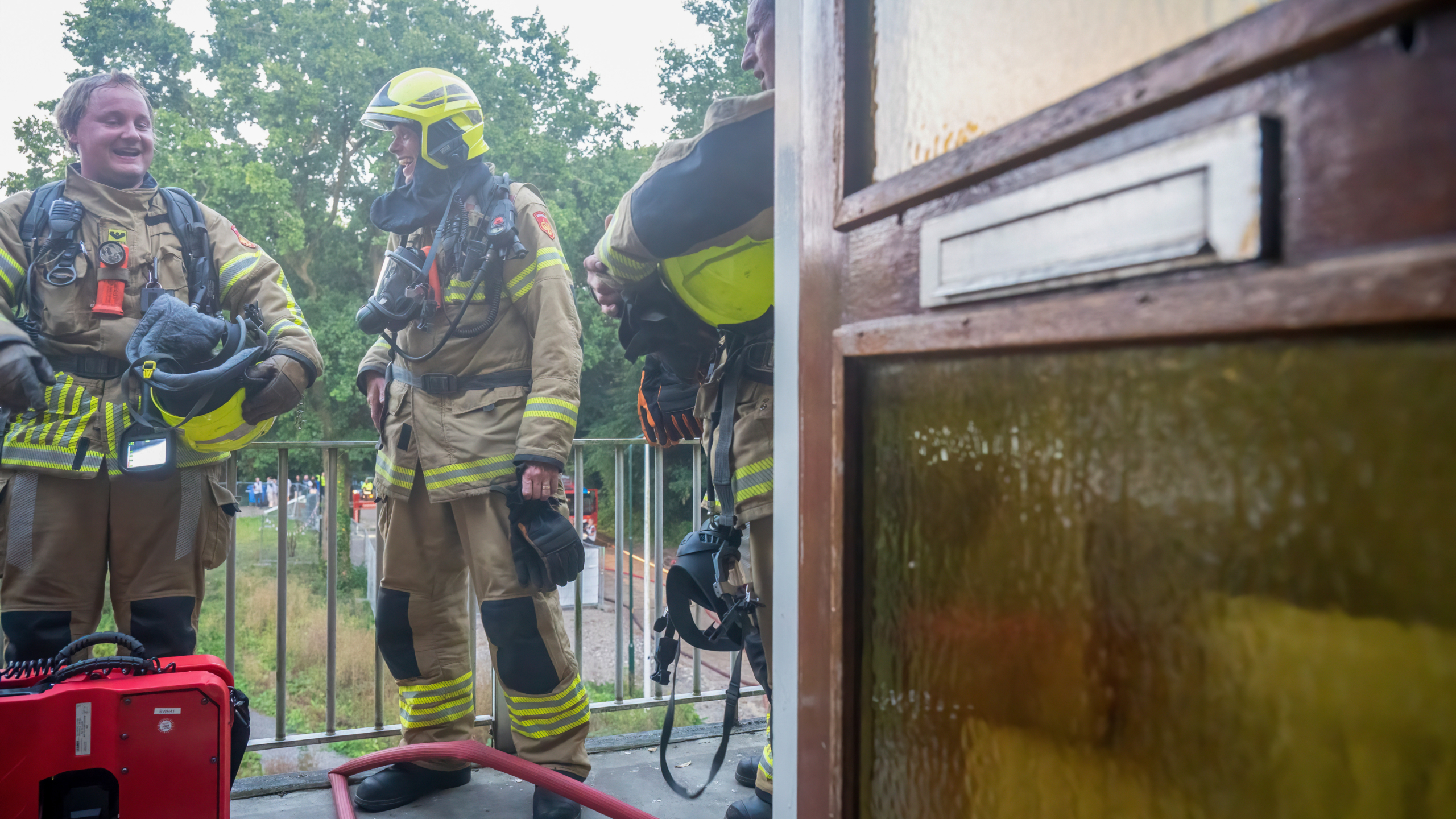 Brandweerlieden in volledige uitrusting staan buiten na een interventie, met hun helmen afgenomen en een brandweerslang op de grond voor hen, naast een houten deur op een balkon.