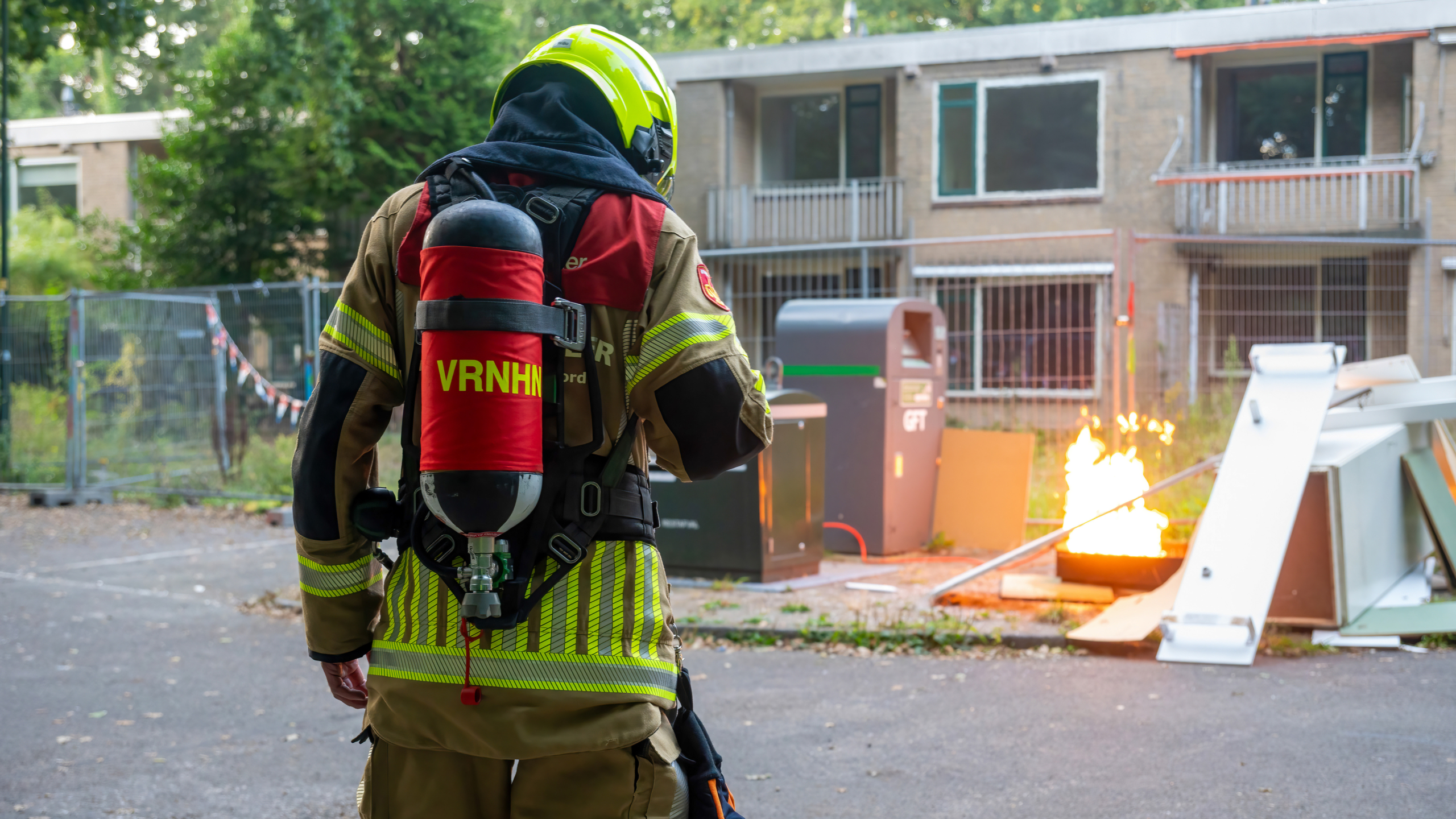 Een brandweerman in uniform en helmmasker, met een rode zuurstoftank op zijn rug, staat op een asfaltweg voor een gebouw terwijl hij naar een kleine brand kijkt.
