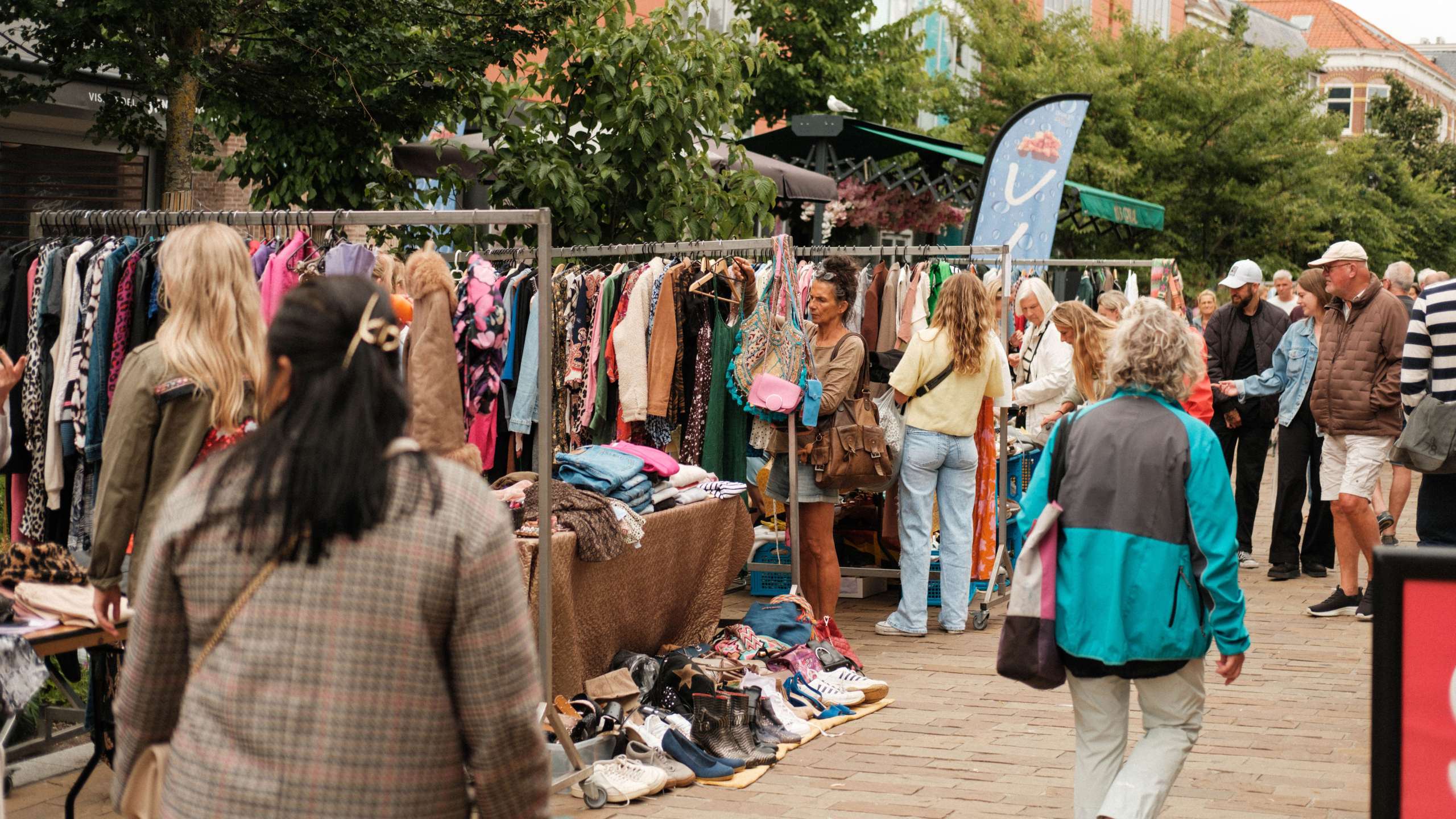 Mensen browsen langs kledingrekken en schoenen op een drukke buitenmarkt, omgeven door bomen en winkels.