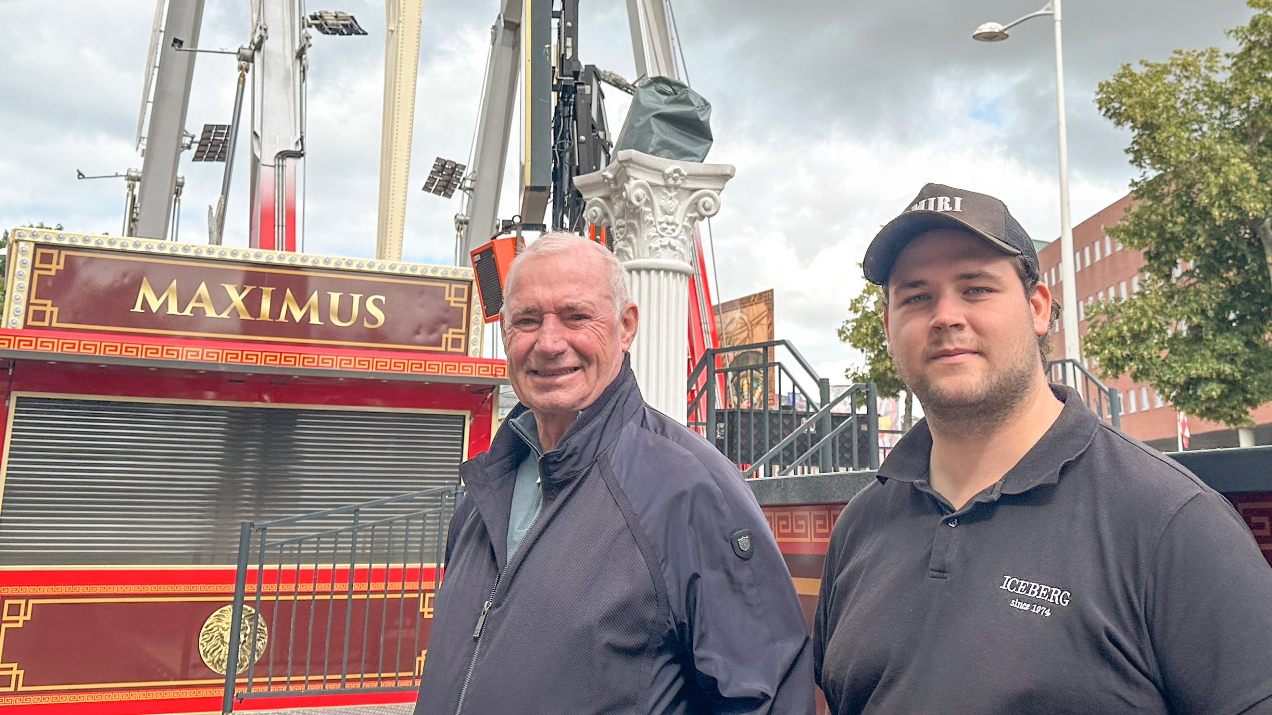 Twee mannen poseren voor een attractie genaamd 'Maximus', met klassieke versieringen en kolommen, op een kermisterrein.