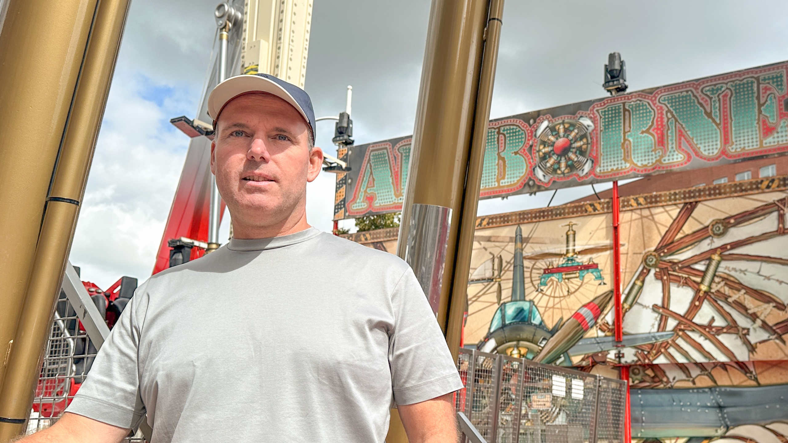 Man in a gray T-shirt and cap standing in front of a carnival ride sign that reads 'AIRBORNE,' with mechanical decorations in the background.