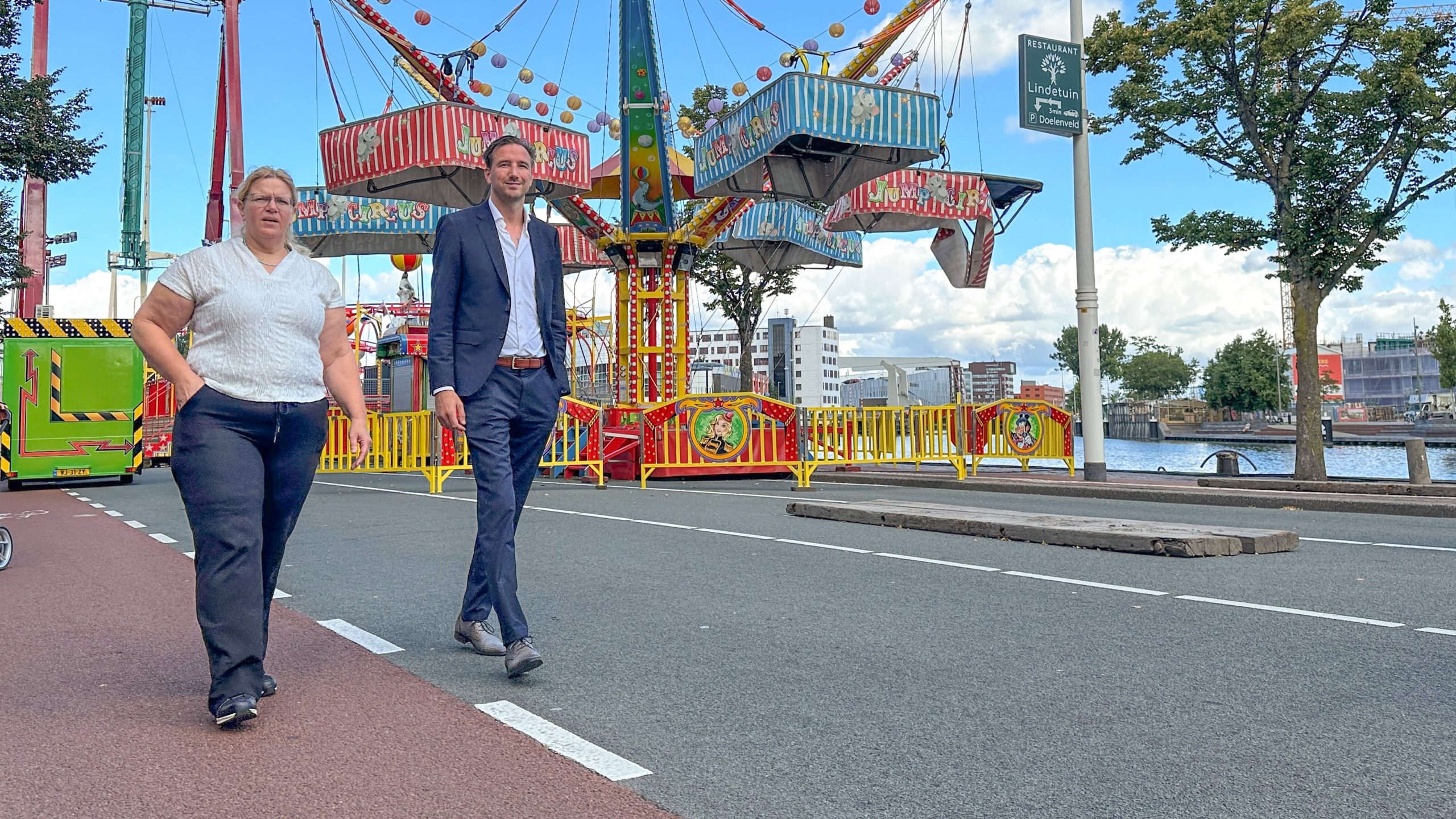 Twee mensen lopen op een trottoir naast een kleurrijke kermisattractie, met een blauwe lucht en water op de achtergrond.