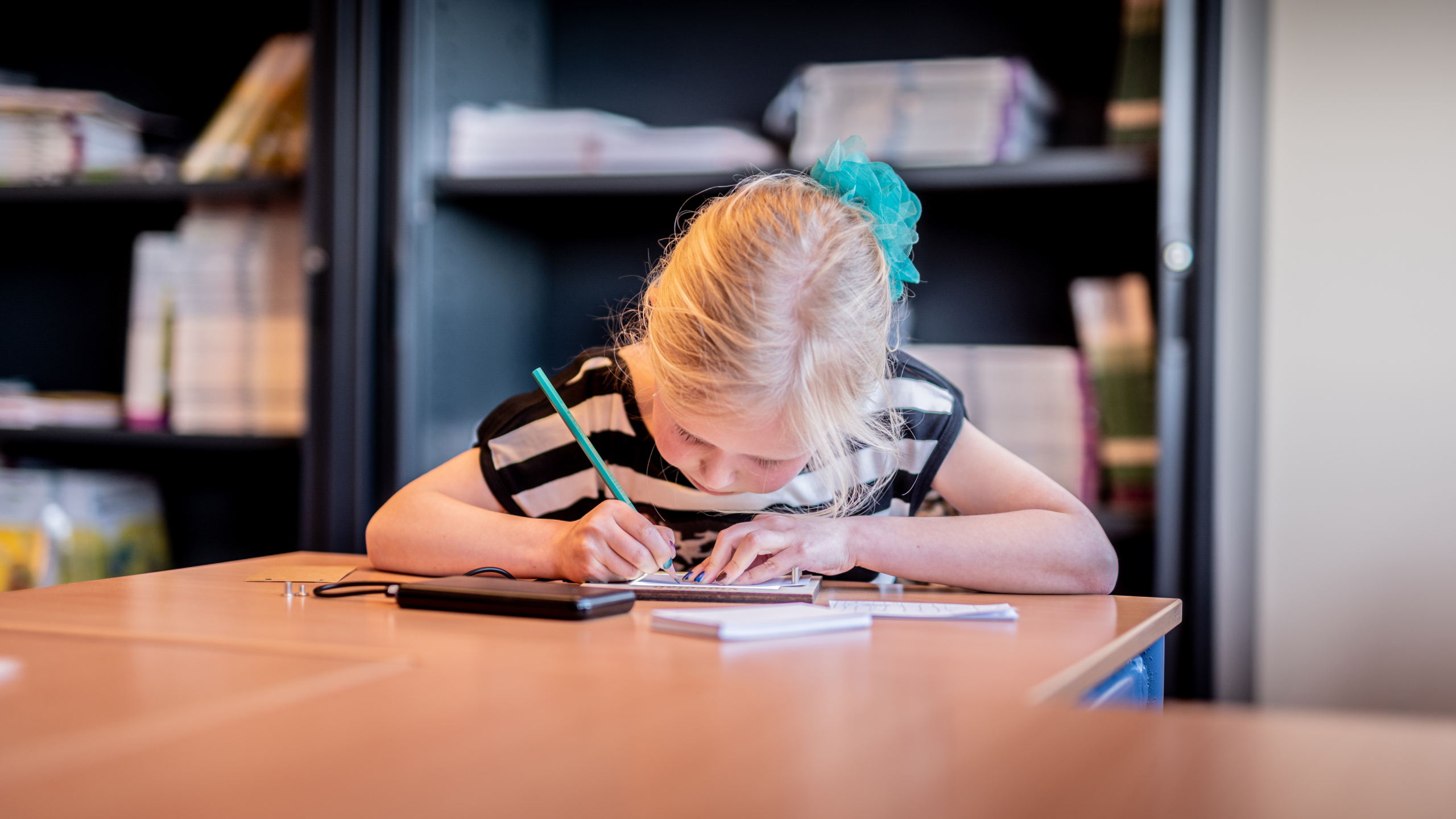 Een jong meisje met een blauw lint in haar haar schrijft geconcentreerd met een potlood in een notitieblok op een tafel, in een ruimte met boekenplanken op de achtergrond.