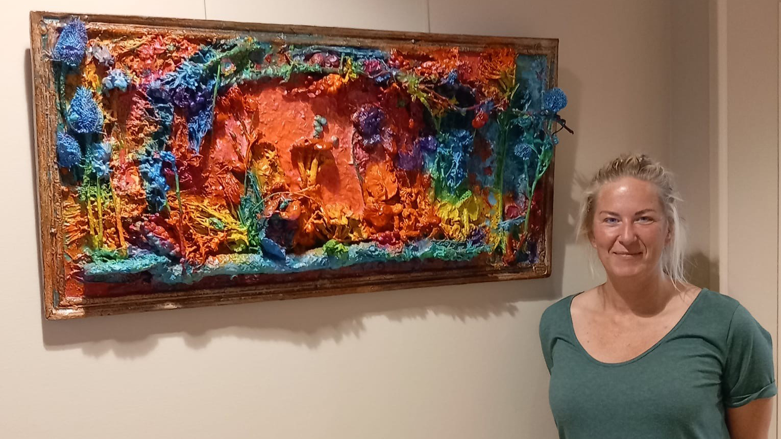 Vrouw in groene trui staat naast een kleurrijk en textuurrijk schilderij met bonte tinten oranje, blauw, geel, groen en rood, geaccentueerd door driedimensionale elementen aan een effen muur.