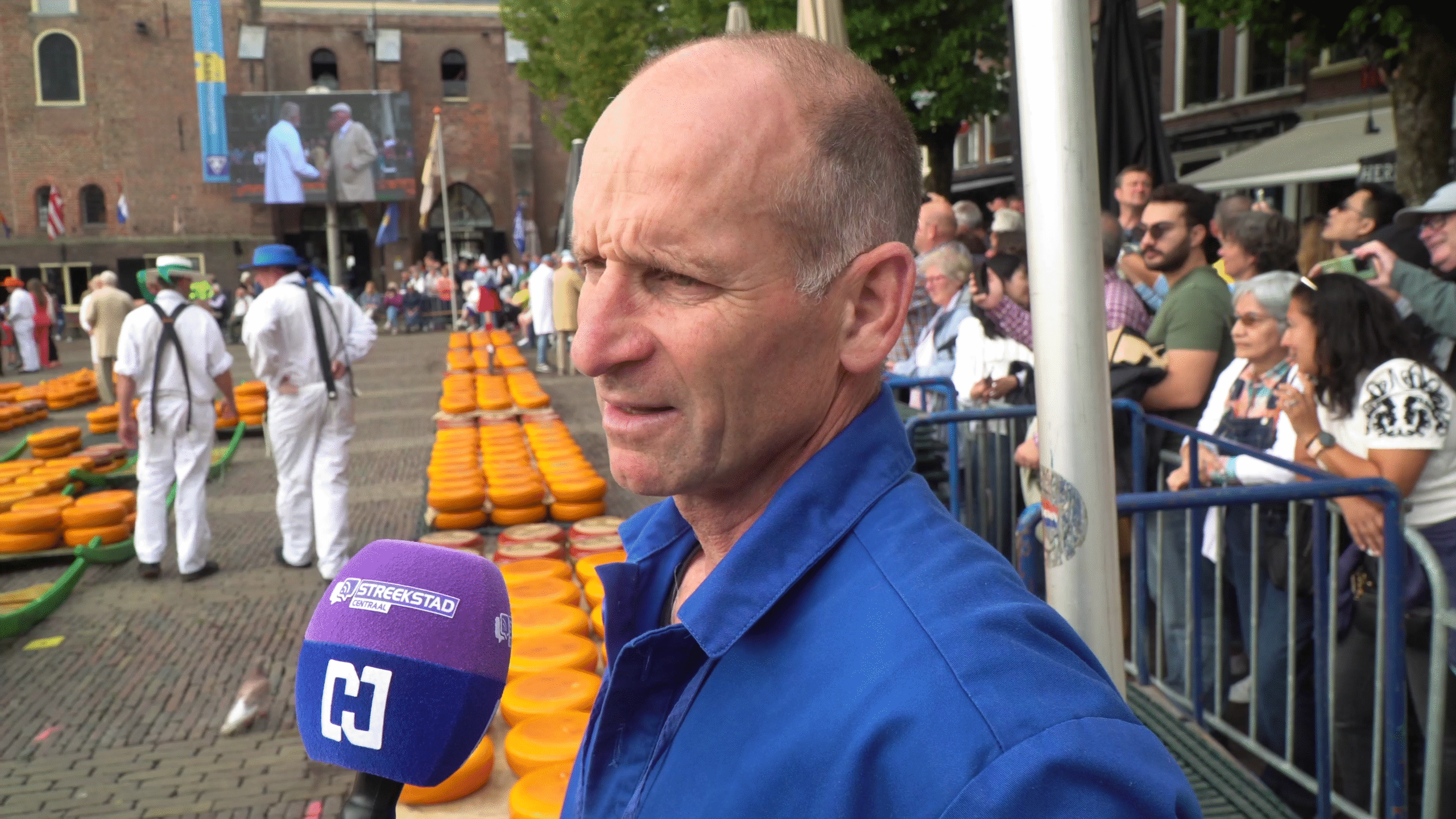 Man in blauwe werkkleding staat voor een rij oranje kazen met microfoon van 'Streekstad Centraal', omringd door een menigte mensen.
