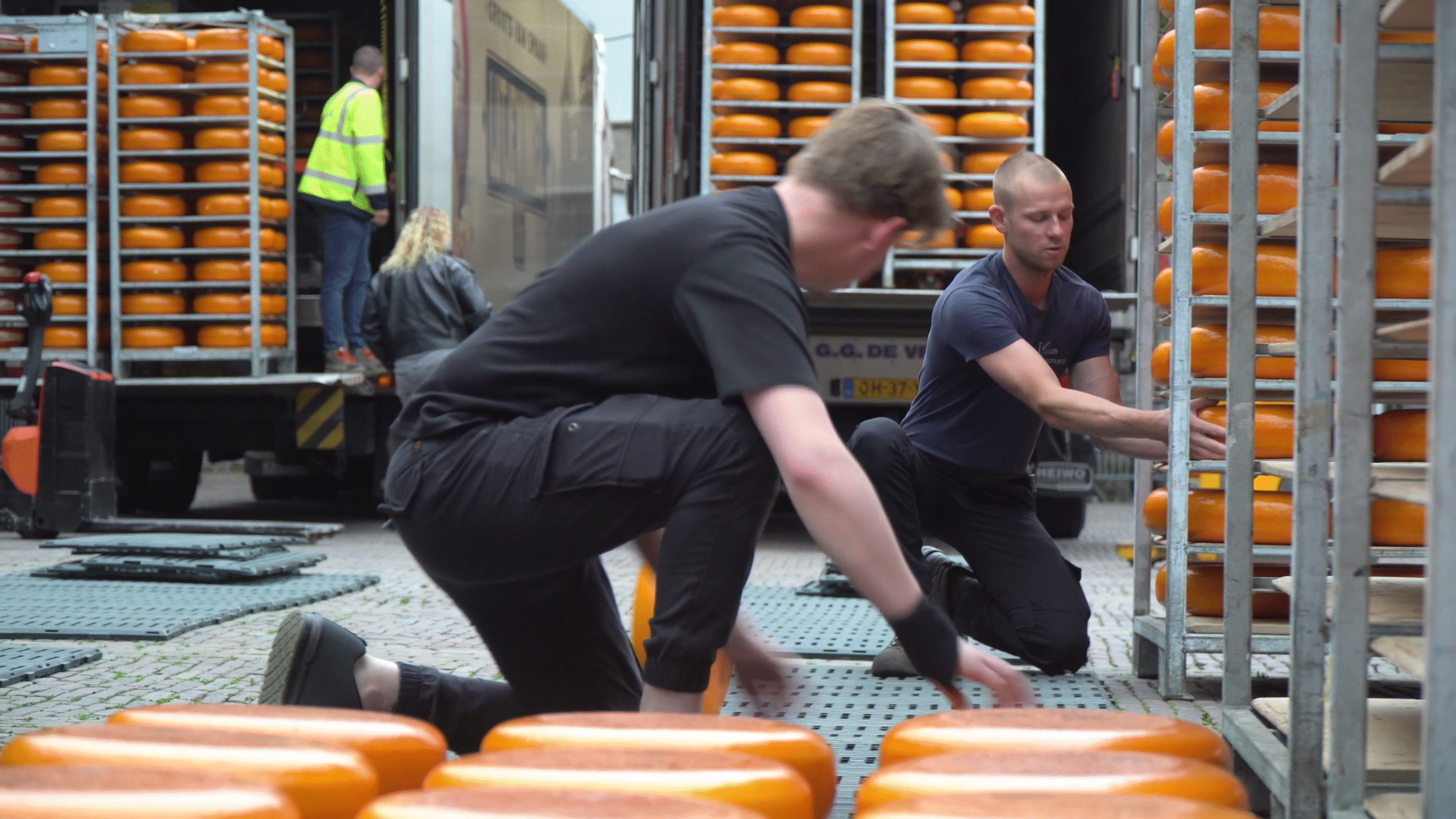 Twee mannen laden grote wielen kaas uit een vrachtwagen op een marktplein.