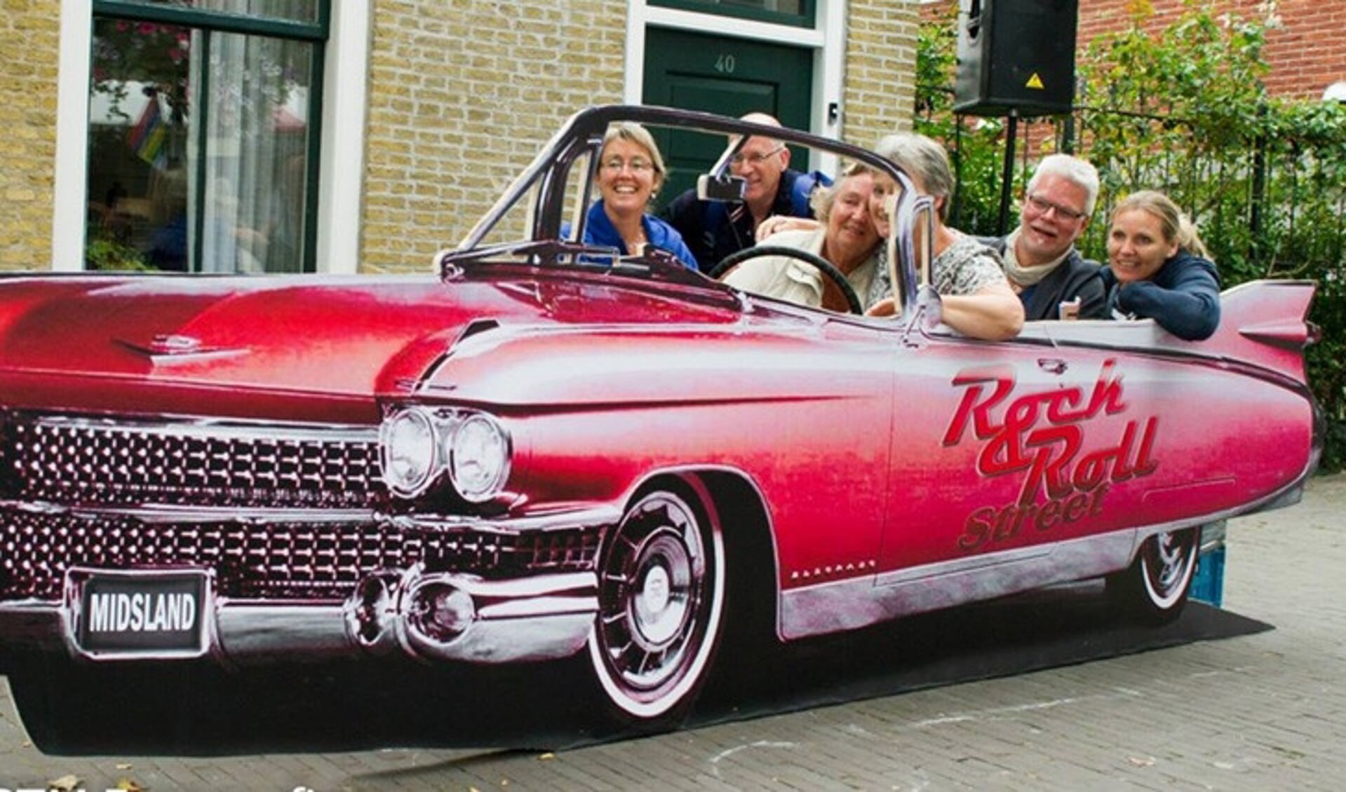 Een groep mensen poseert achter een kartonnen decorstuk van een felrode Cadillac met de tekst 'Rock & Roll street' erop, voor een bakstenen gebouw.