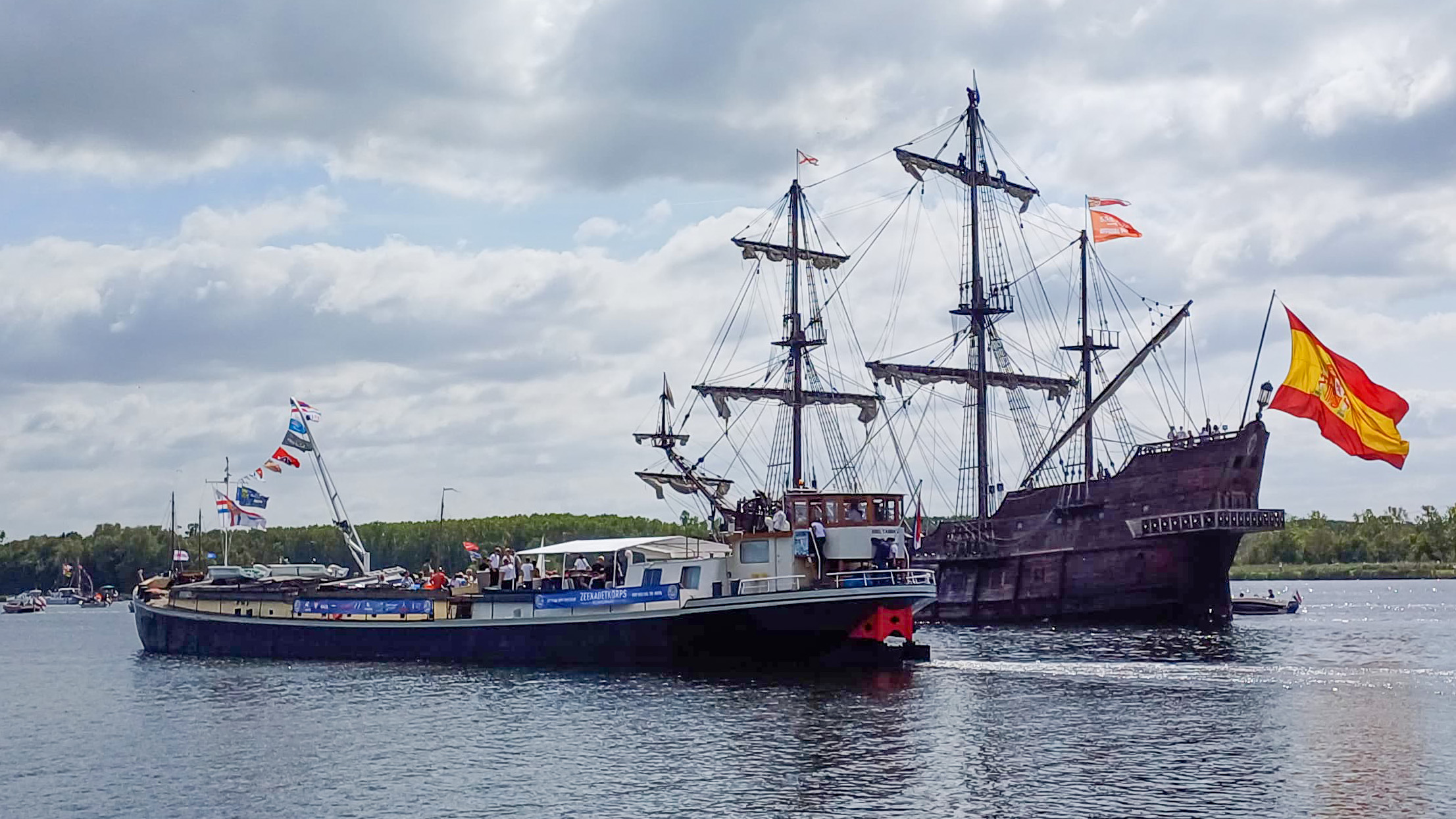 Twee zeilschepen varen zij aan zij, met een groot houten schip met een Spaanse vlag en een kleiner modern schip met kleurrijke vlaggen, onder een bewolkte hemel.