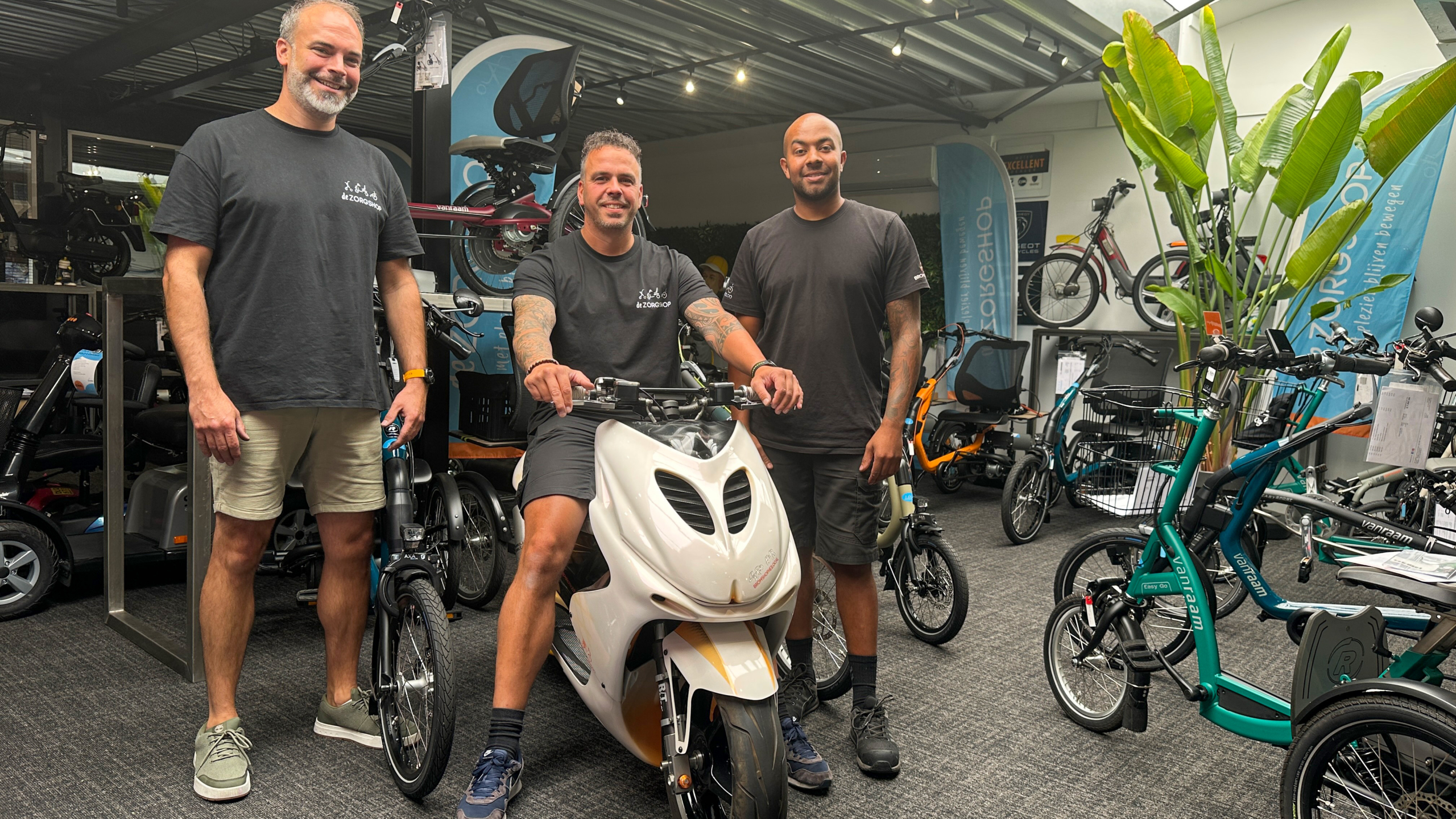 Drie mannen staan in een winkel met scooters en fietsen, waarvan een man op een witte scooter zit. Ze dragen casual kleding en kijken lachend naar de camera.