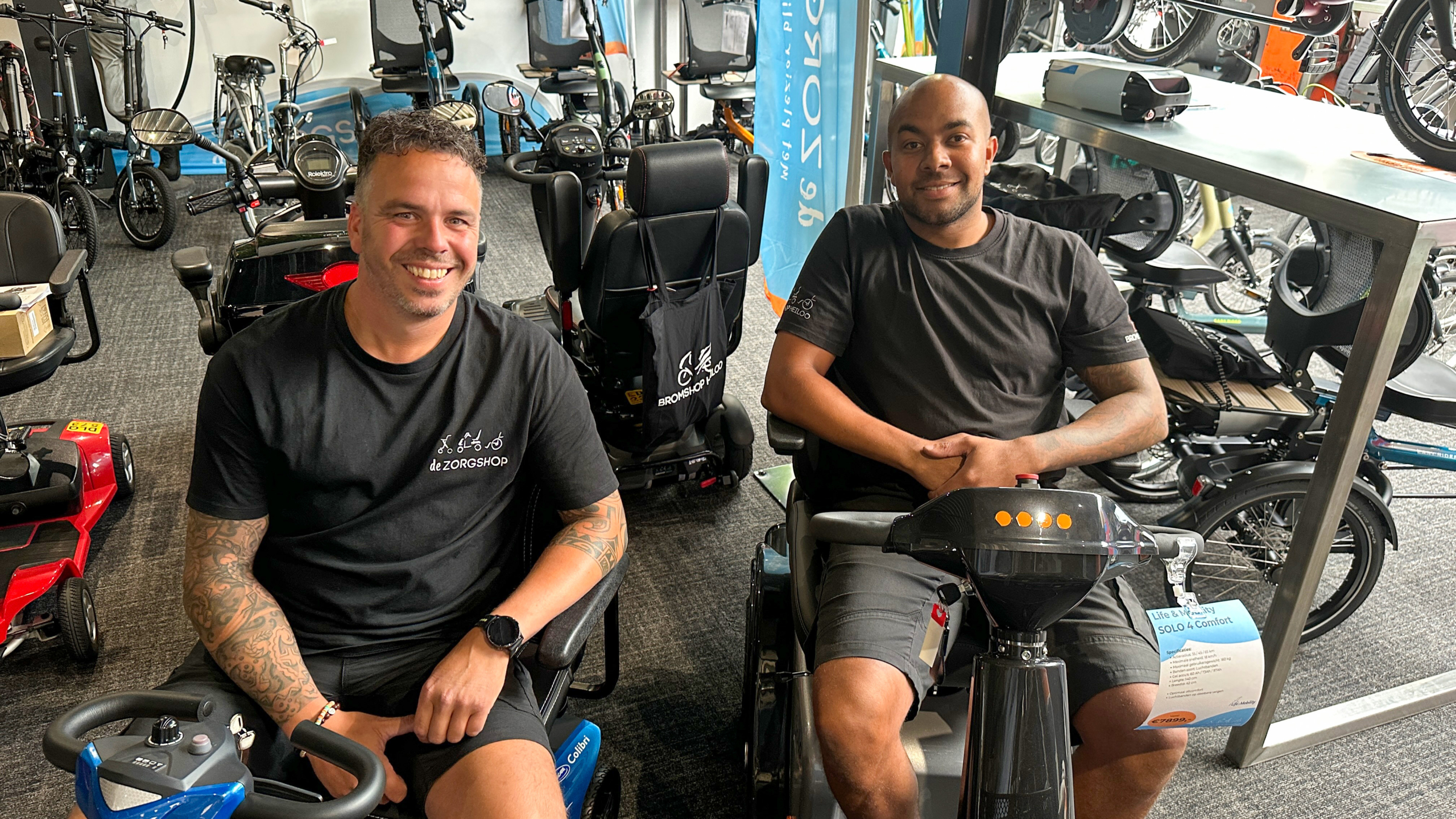 Twee mannen zitten lachend op scootmobiels in een winkel, omgeven door diverse mobiliteitshulpmiddelen en fietsen, terwijl ze kleding dragen met het logo van 'de ZORGSHOP'.