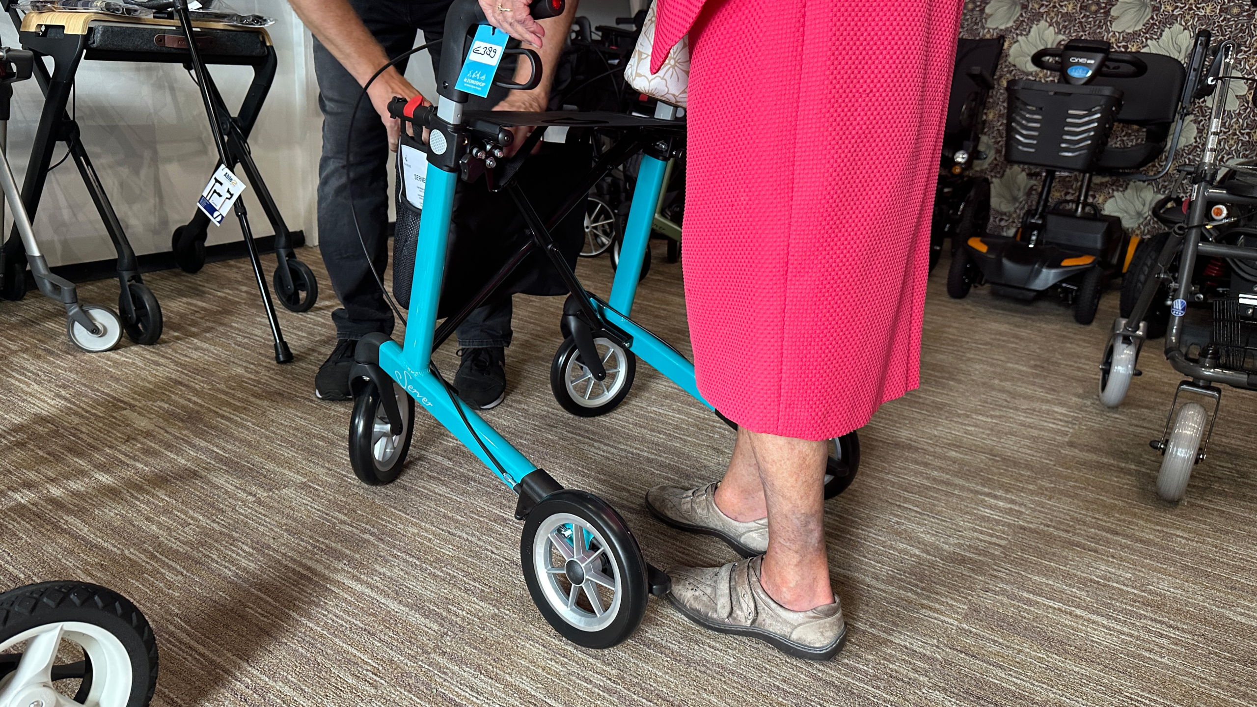 Een persoon in een roze kledingstuk staat naast een turquoise rollator, terwijl een andere persoon hen helpt, in een winkel met verschillende soorten rollators en scootmobielen op de achtergrond.
