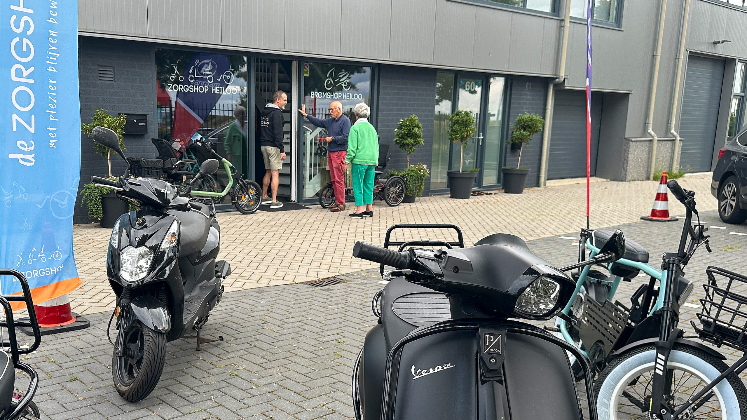 Mensen staan buiten bij een winkel met fietsen en scooters geparkeerd op de voorgrond.