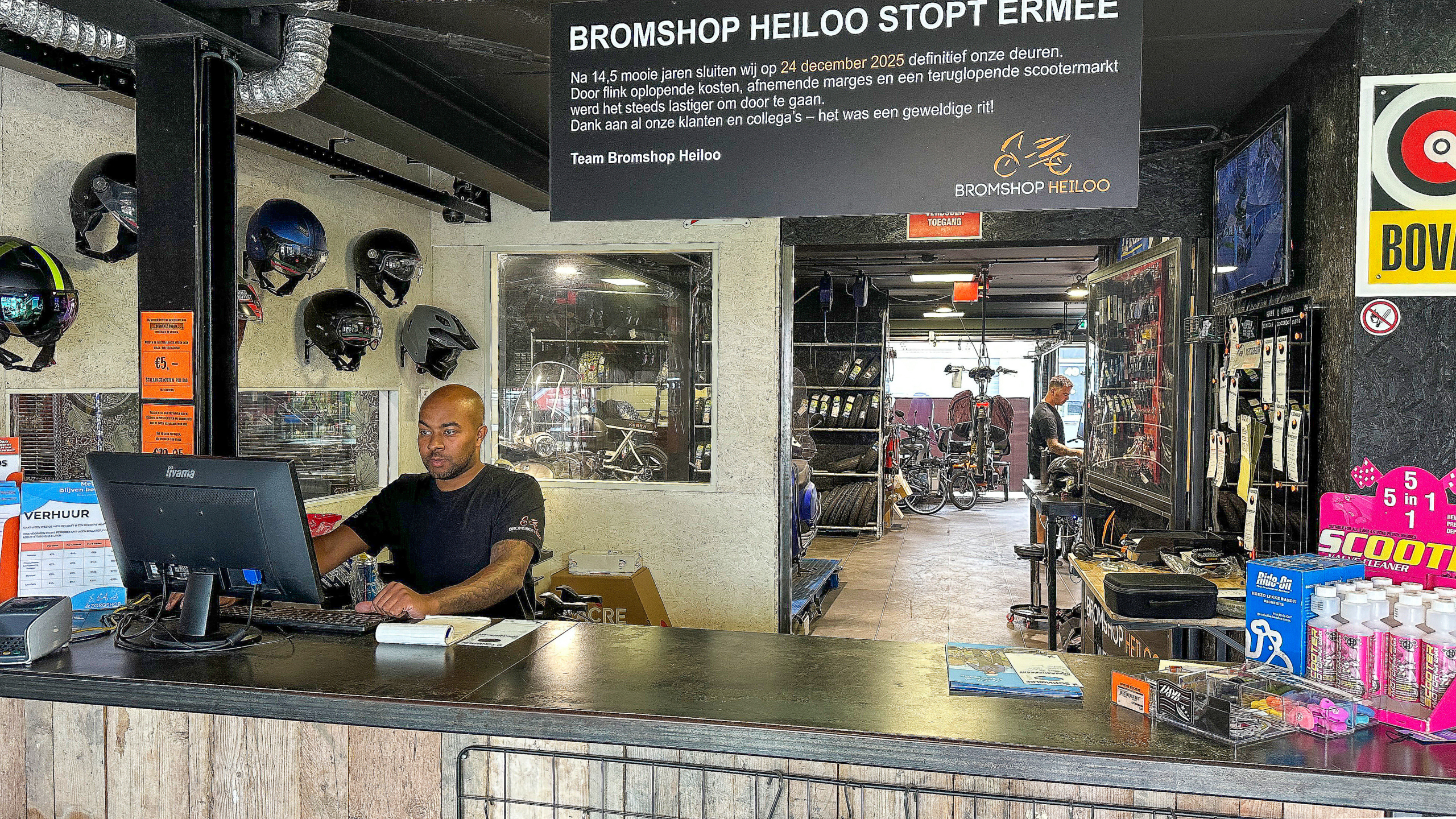 Man achter de balie in een scooterwinkel, met helmen aan de muur en een bord met de tekst over de sluiting van de winkel Bromshop Heiloo.