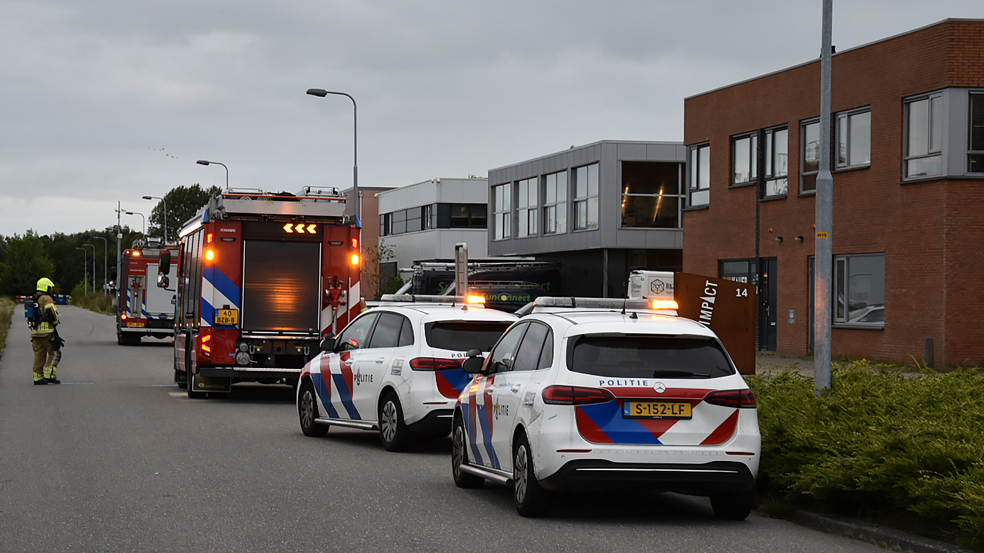 Brandweer- en politiewagens staan geparkeerd op een industrieterrein, met een brandweerman in uniform lopend naar een gebouw.