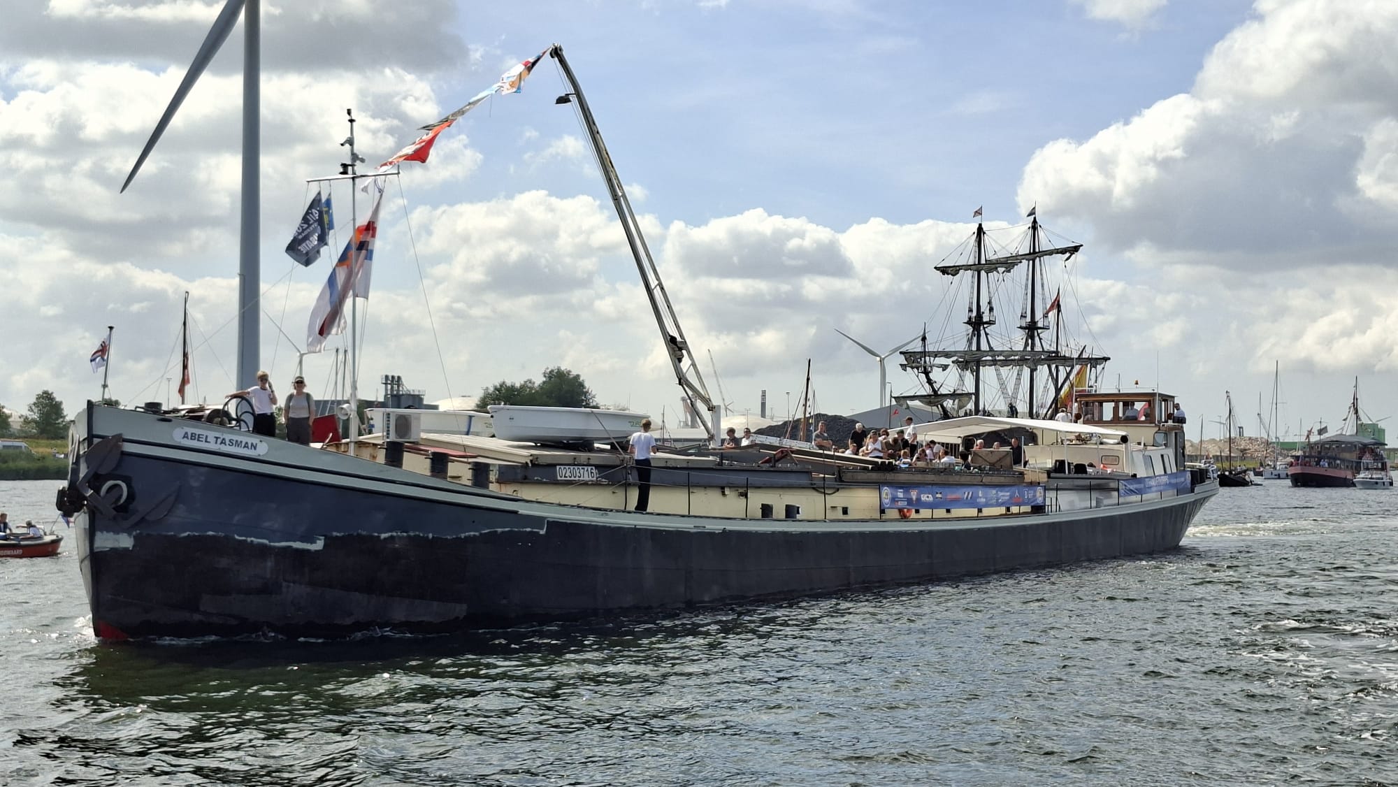 Groot schip met de naam "Abel Tasman" vaart op het water, omringd door mensen aan boord en met een windmolen op de achtergrond.