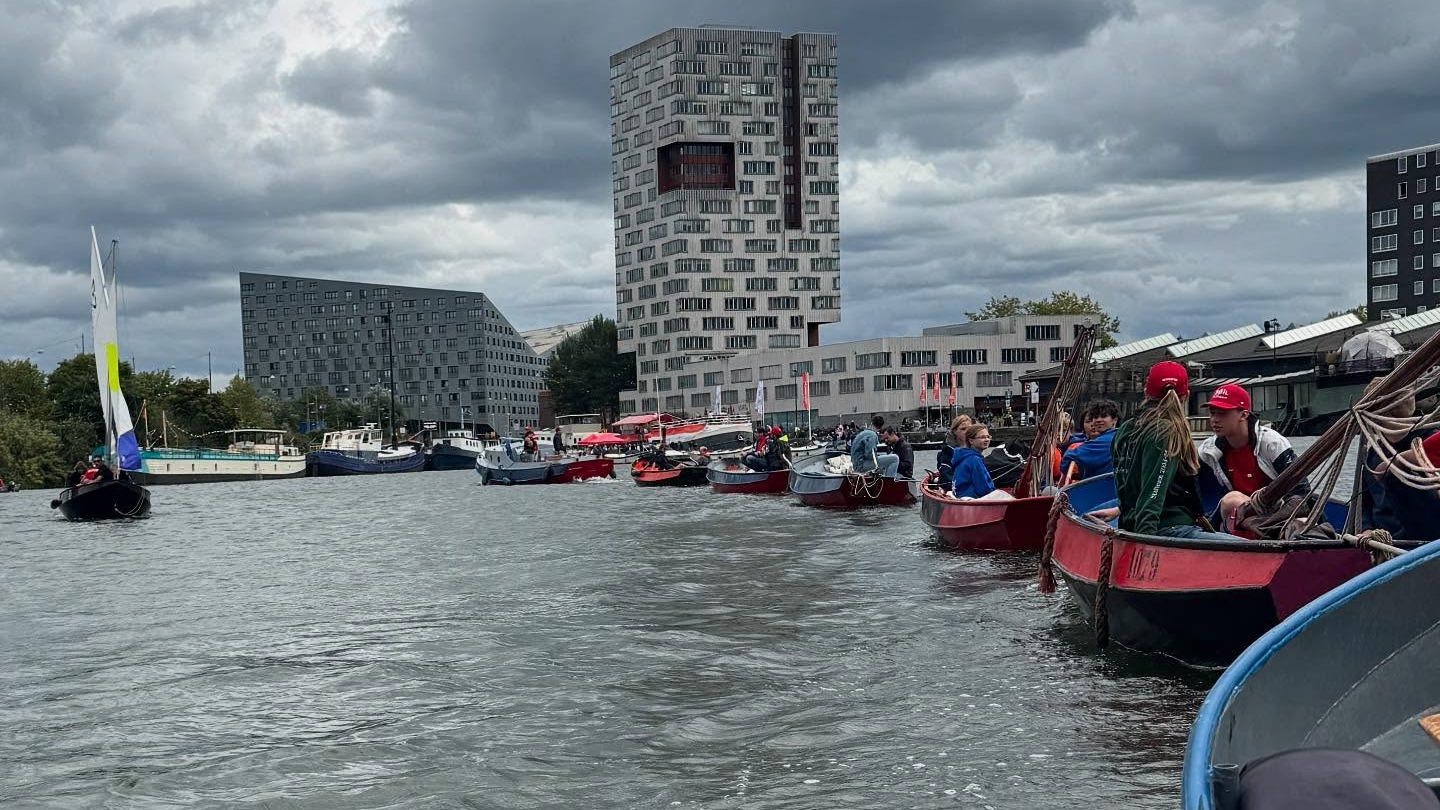 Een groep mensen vaart in kleine boten op een rivier in een stedelijke omgeving, met moderne gebouwen op de achtergrond onder een bewolkte hemel.