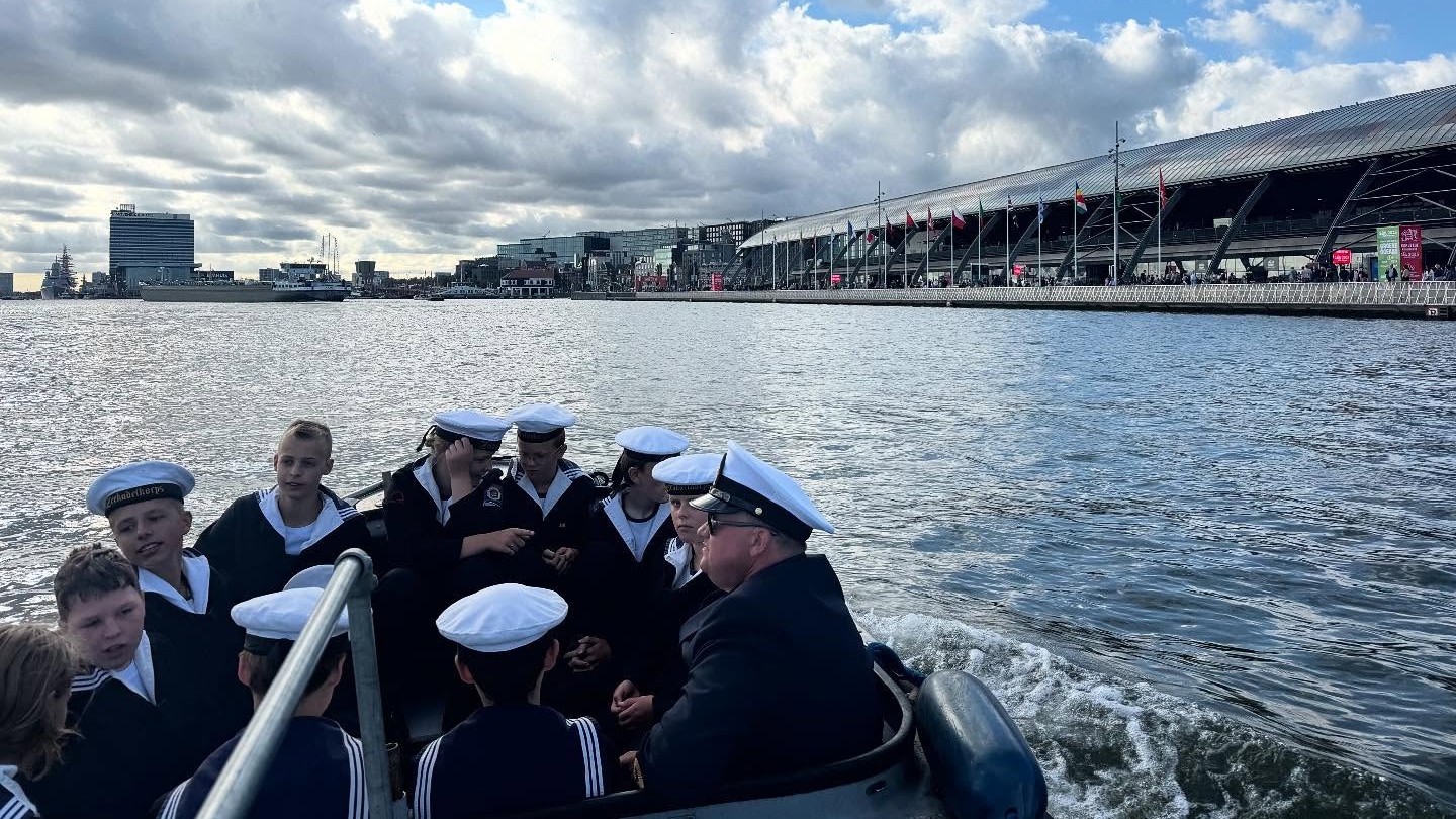Kleine groep zeekadetten in marine-uniformen met witte petten zitten in een boot op het water, met een grote overdekte terminal rechts en gebouwen aan de horizon onder een bewolkte lucht.