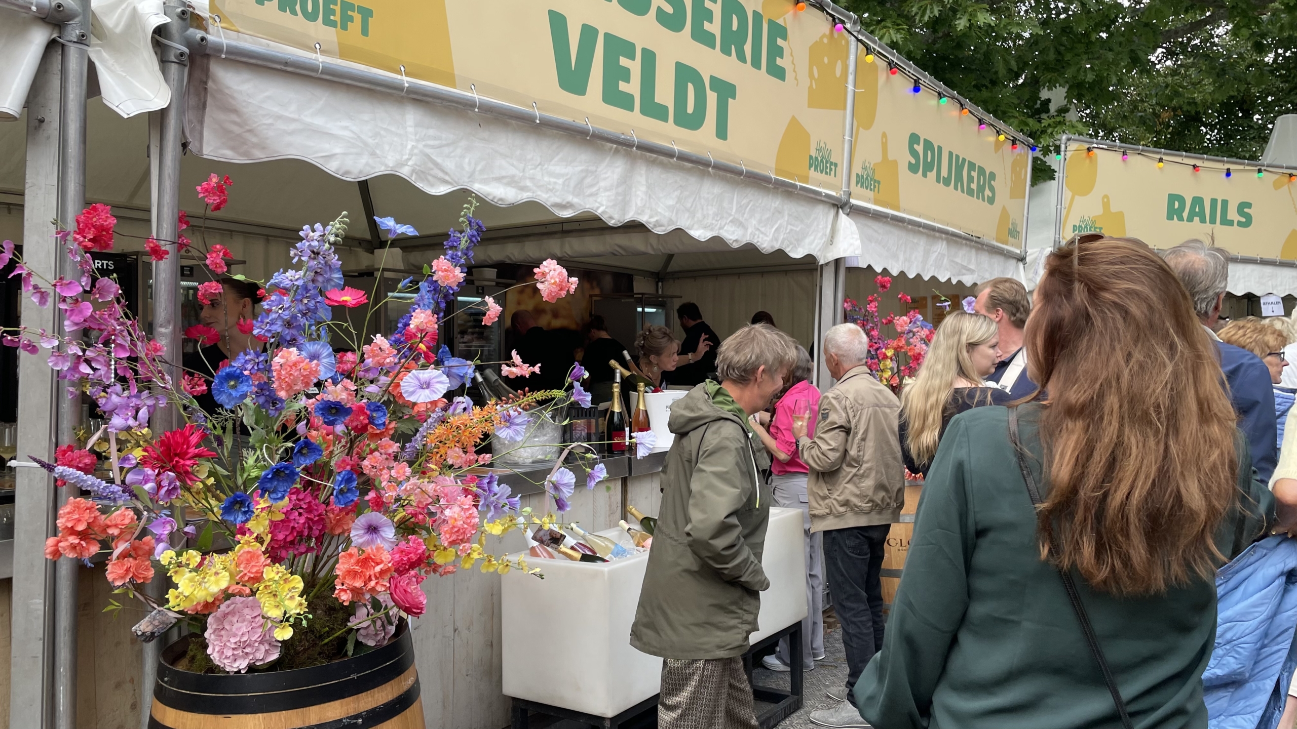 Groep mensen in de rij bij een etenskraam op een buitenfestival, versierd met kleurrijke bloemen en flessen drank in ijs.