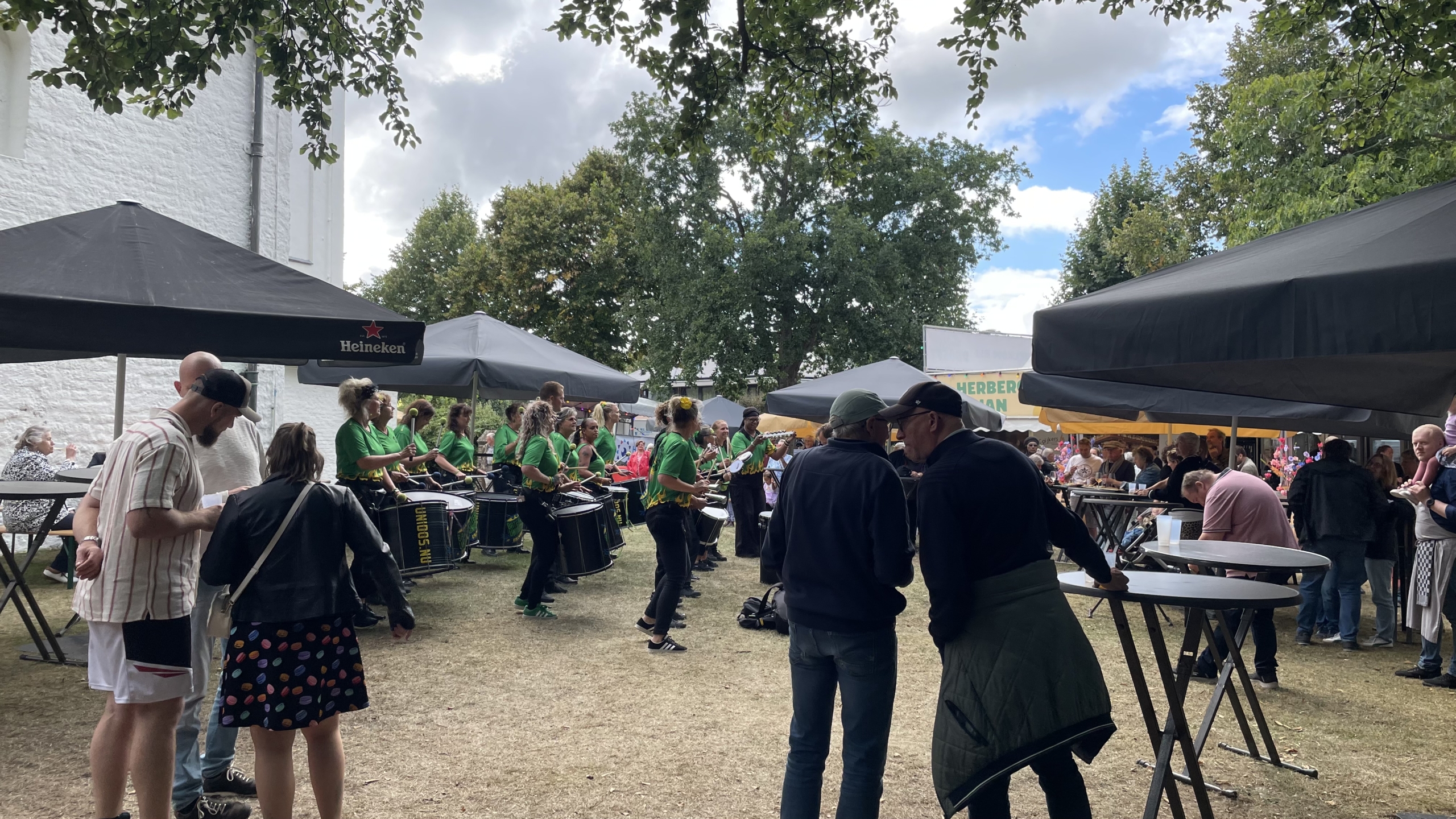Een groep mensen verzameld onder partytenten tijdens een buitenevenement, met muzikanten in groene shirts die spelen op instrumenten. Er zijn tafels en mensen aan het praten terwijl anderen toekijken.