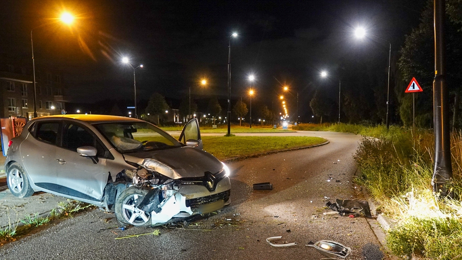 Auto met aanzienlijke schade aan de voorzijde na een ongeval, geparkeerd op een weg bij nachtelijke straatverlichting, met verspreide brokstukken op het asfalt en een beschadigde lantaarnpaal recht vooruit.